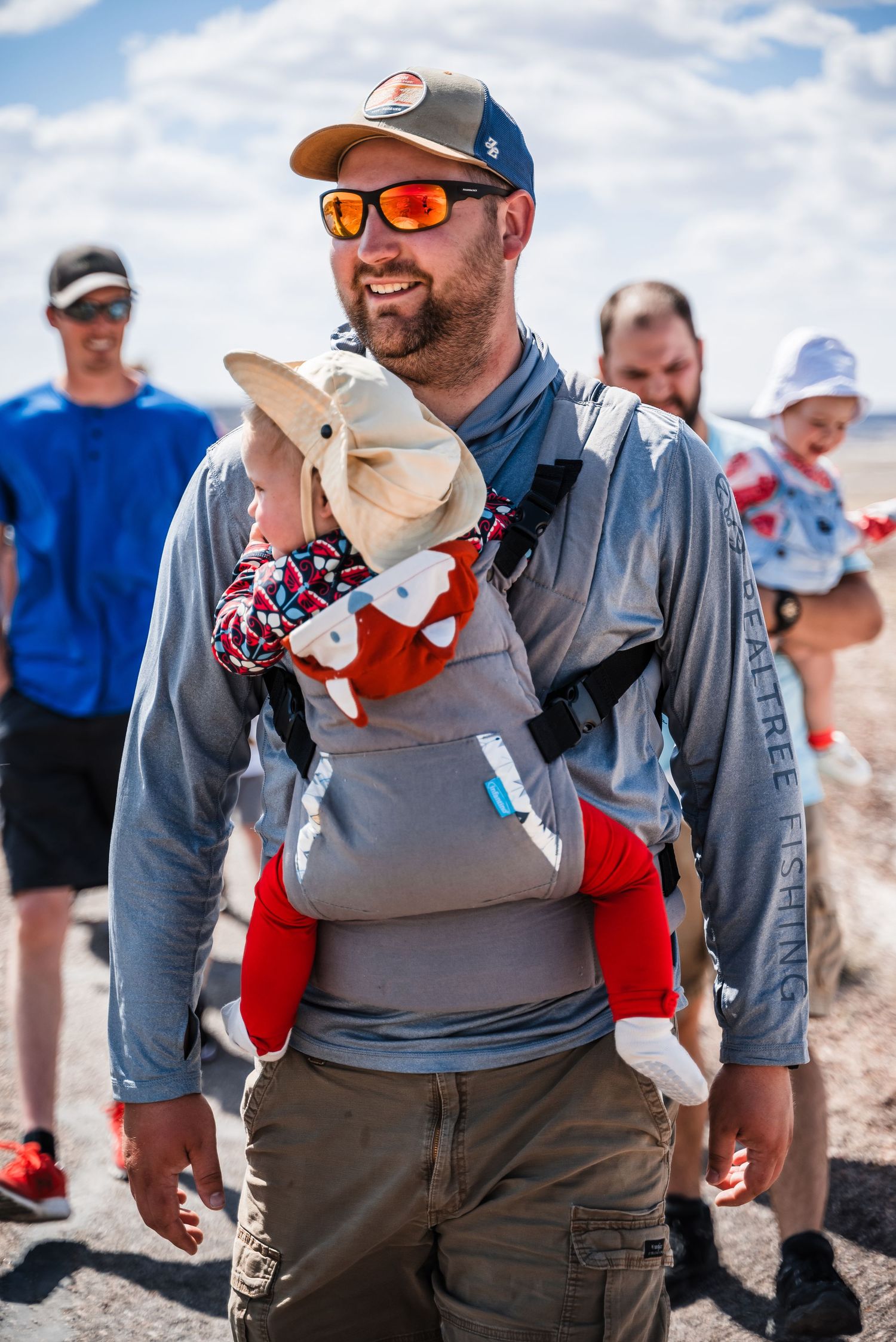 Hiking outdoors in desert terrain wearing sunglasses and carrying a small companion in a baby carrier.
