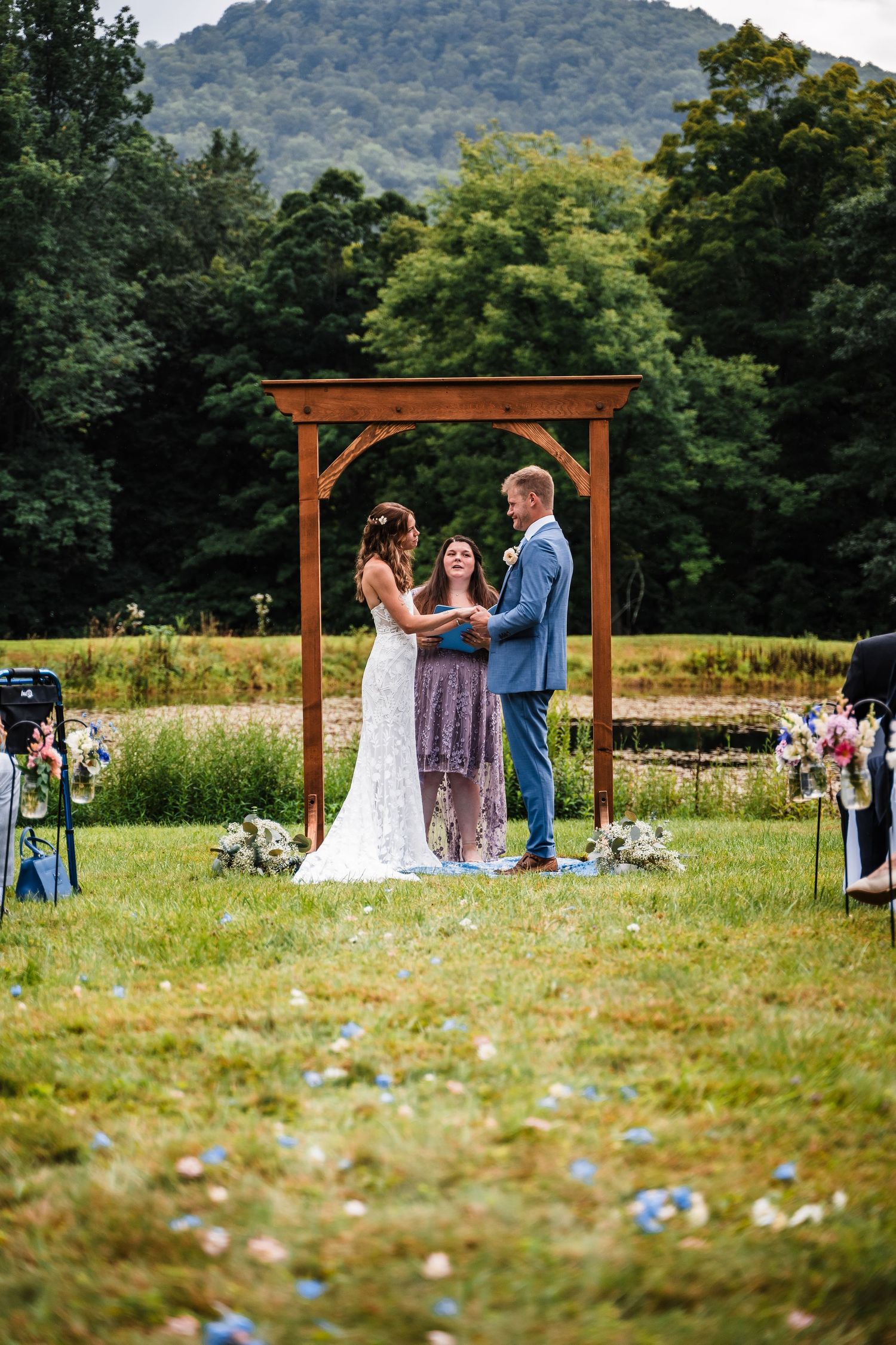 An intimate wedding ceremony takes place under a wooden arch with mountains in the background.