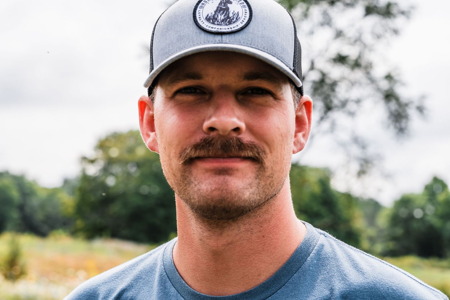 Person wearing a gray trucker hat and blue t-shirt smiling at the camera outdoors.