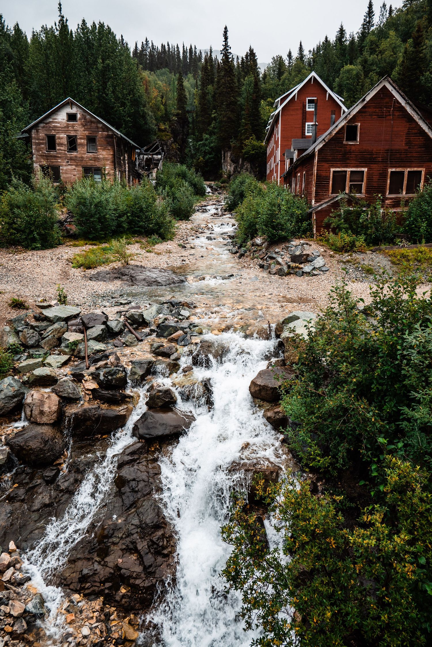Historic red wooden buildings perched along a rushing mountain stream in a forest setting.