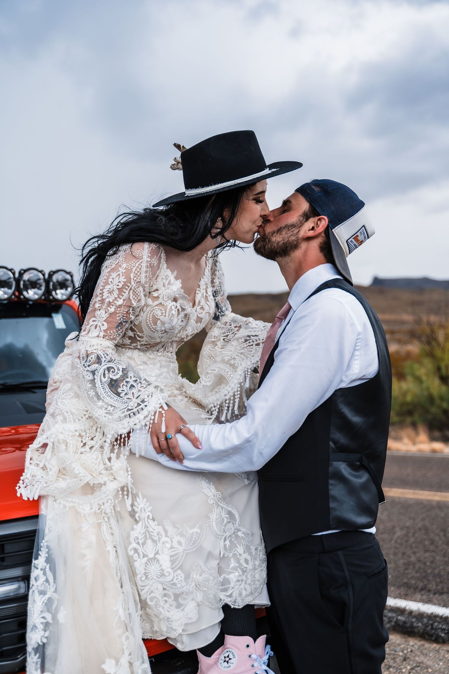 Wedding couple in rustic attire embracing near vintage red vehicle in desert landscape.