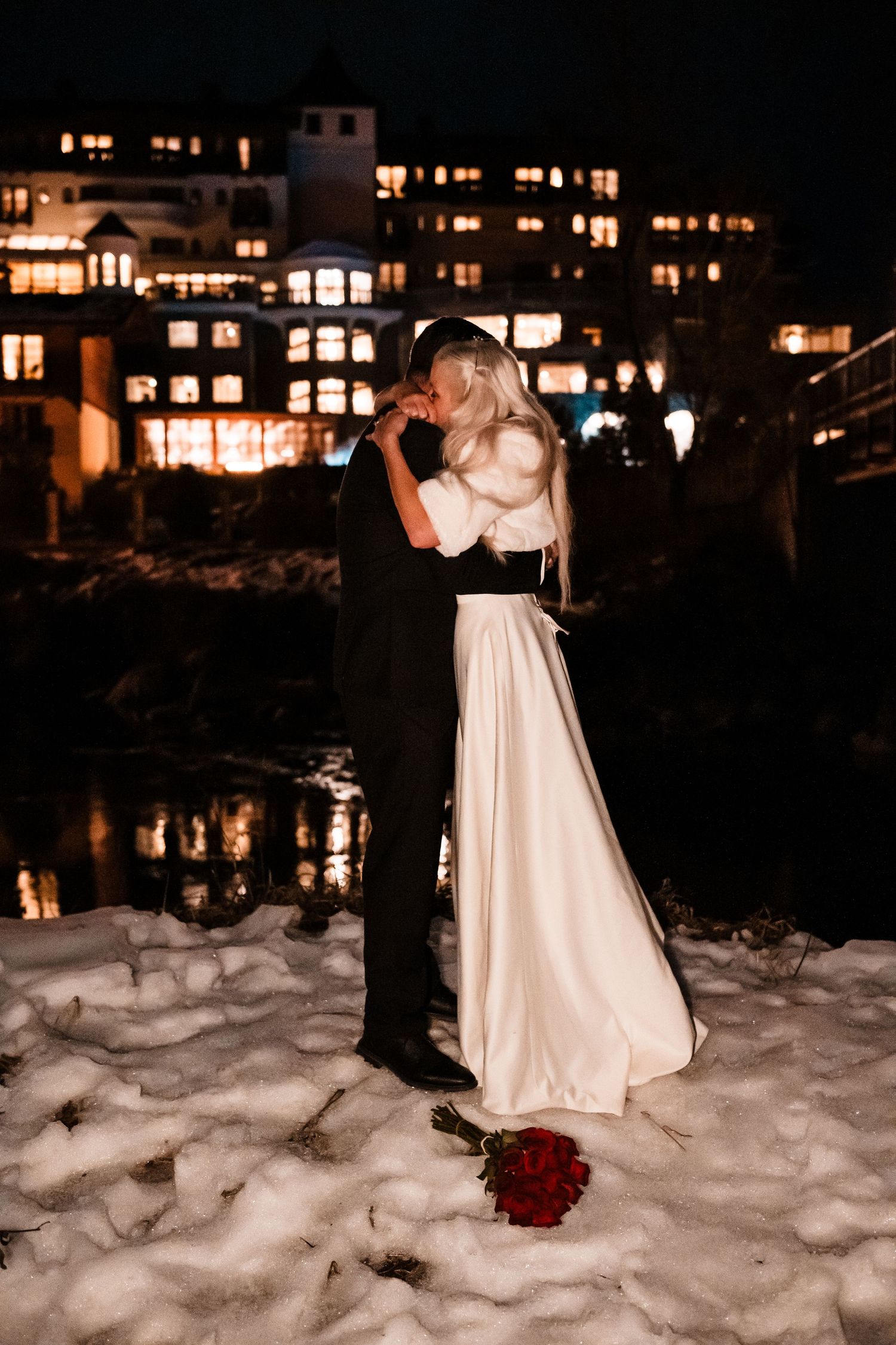 A winter wedding photo at night with city lights and snow, featuring a red rose bouquet.