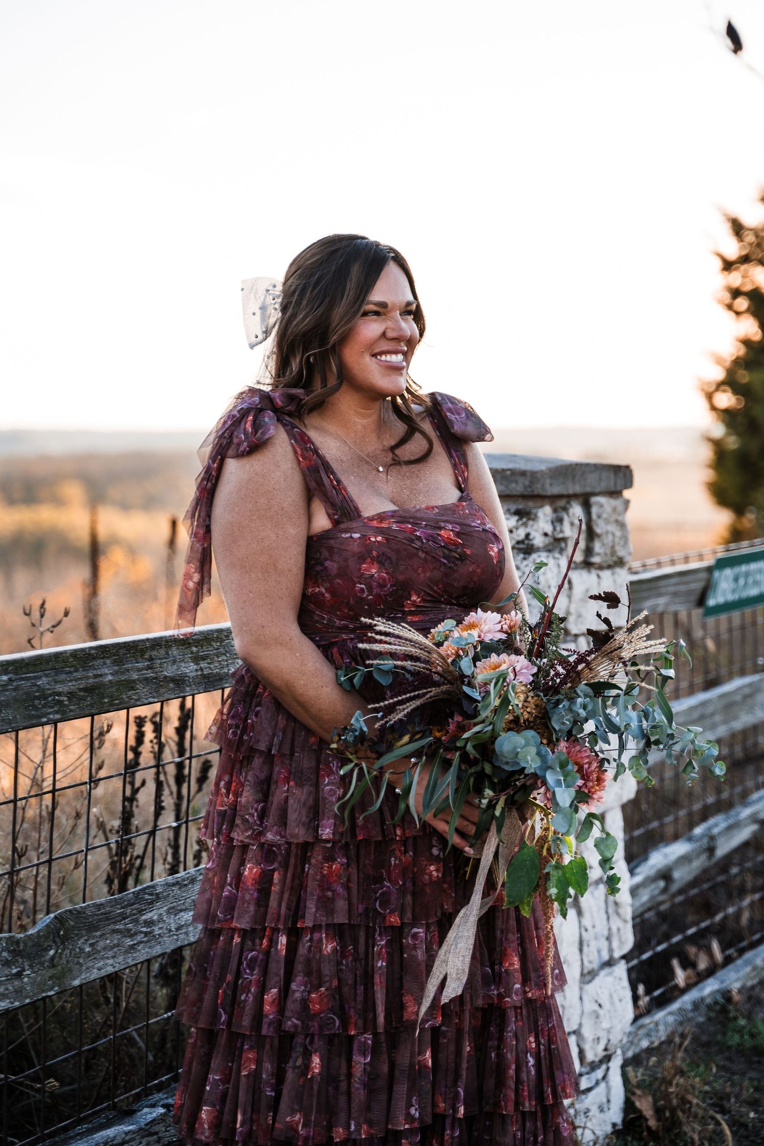 Person in a burgundy tiered dress holds a floral bouquet against a rustic stone wall backdrop.