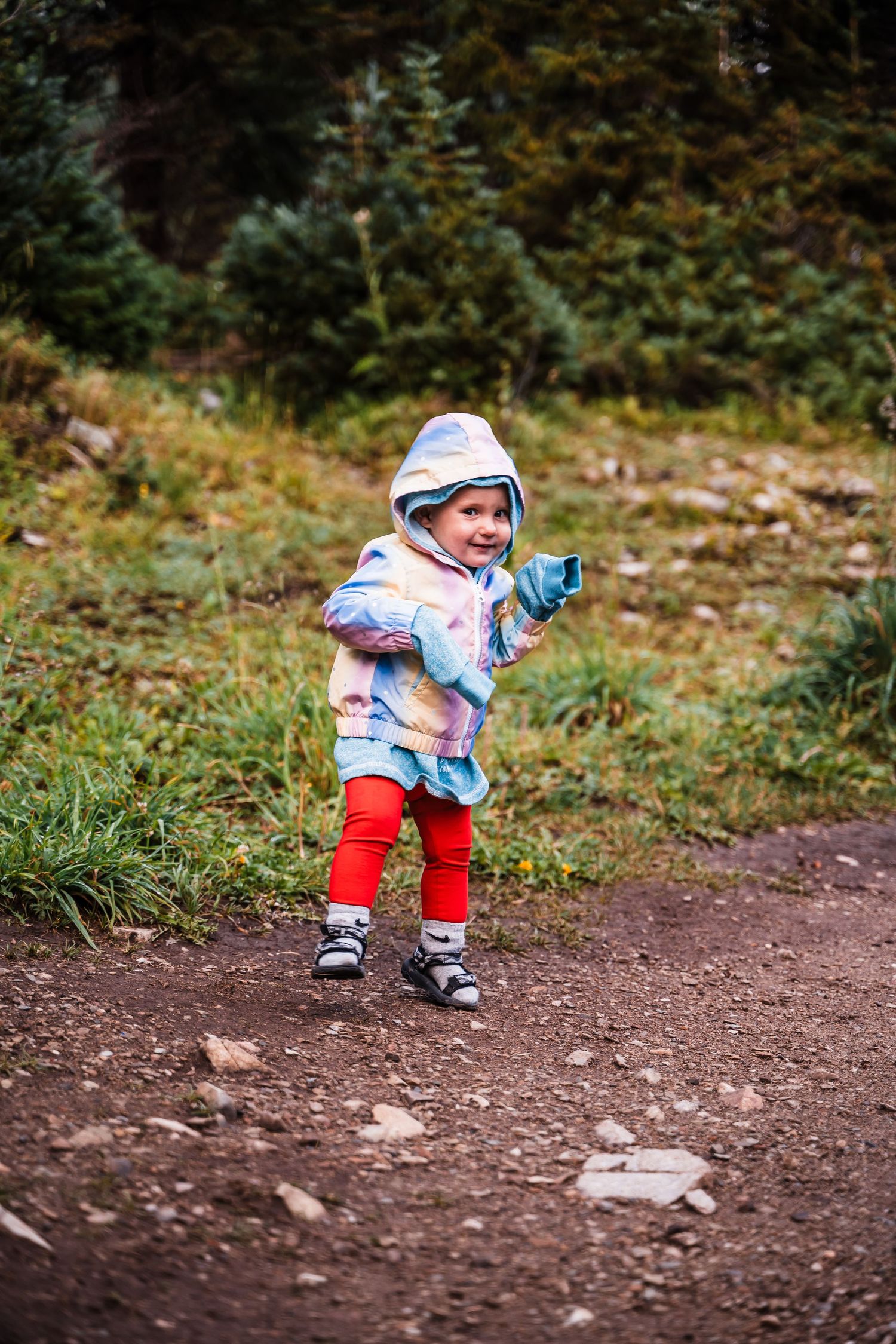 A small child in winter clothing plays joyfully on a dirt trail surrounded by forest scenery.