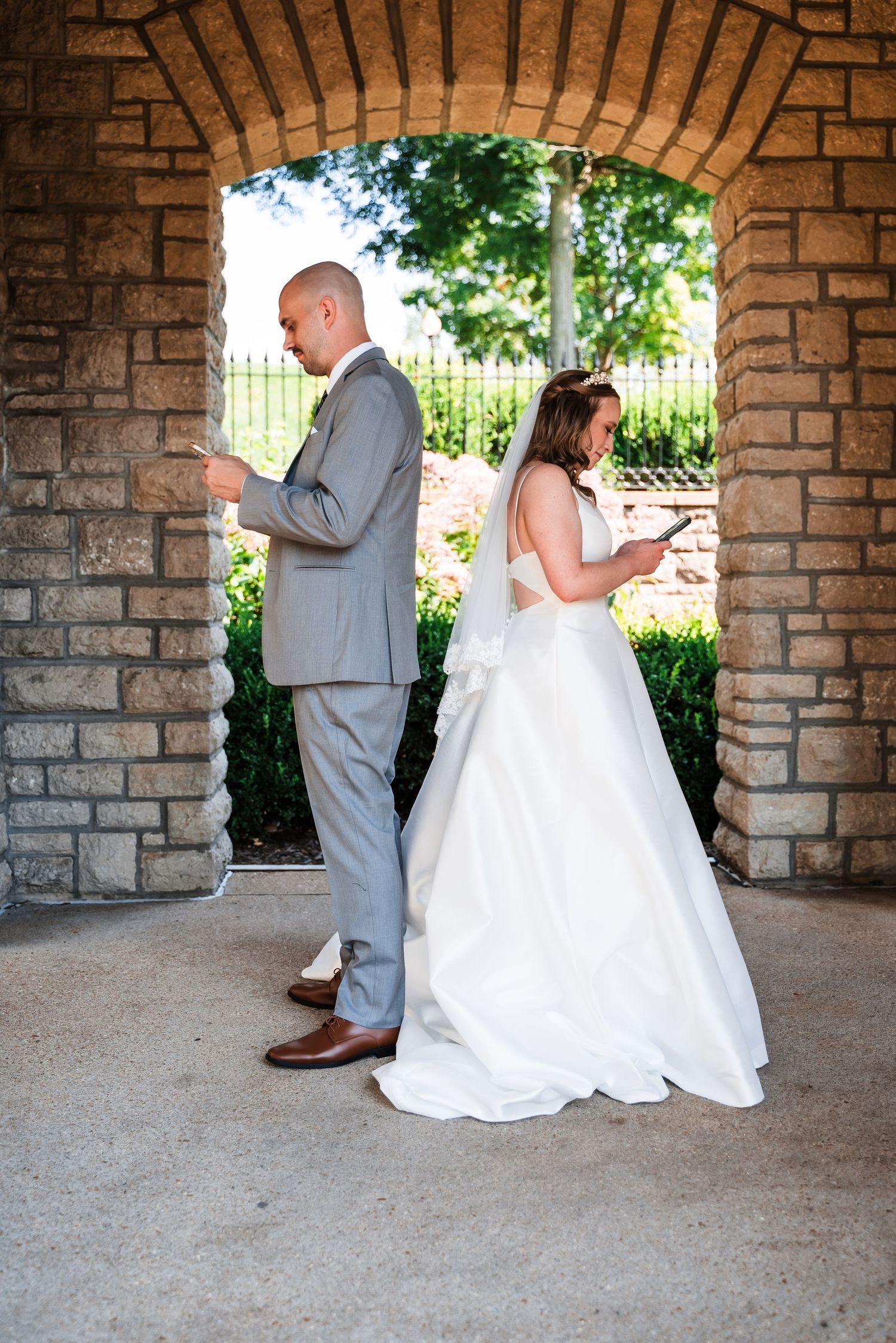 A couple stands back to back reading private notes before their wedding ceremony under a stone archway.