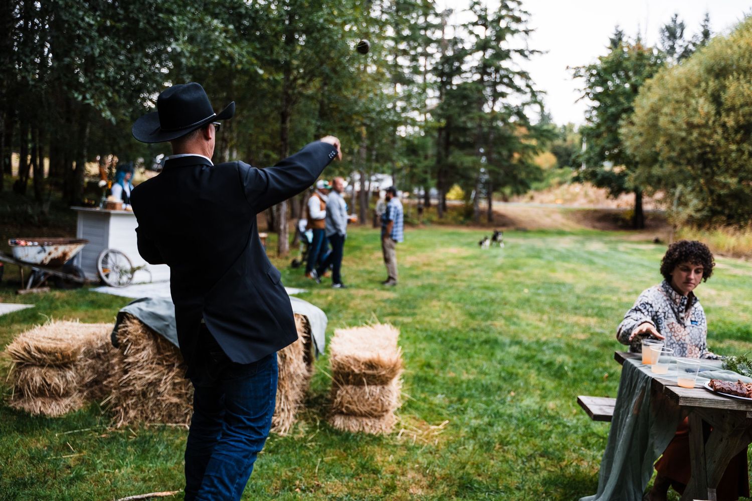 People enjoying outdoor activities with hay bales and tables set up in a grassy field surrounded by trees.