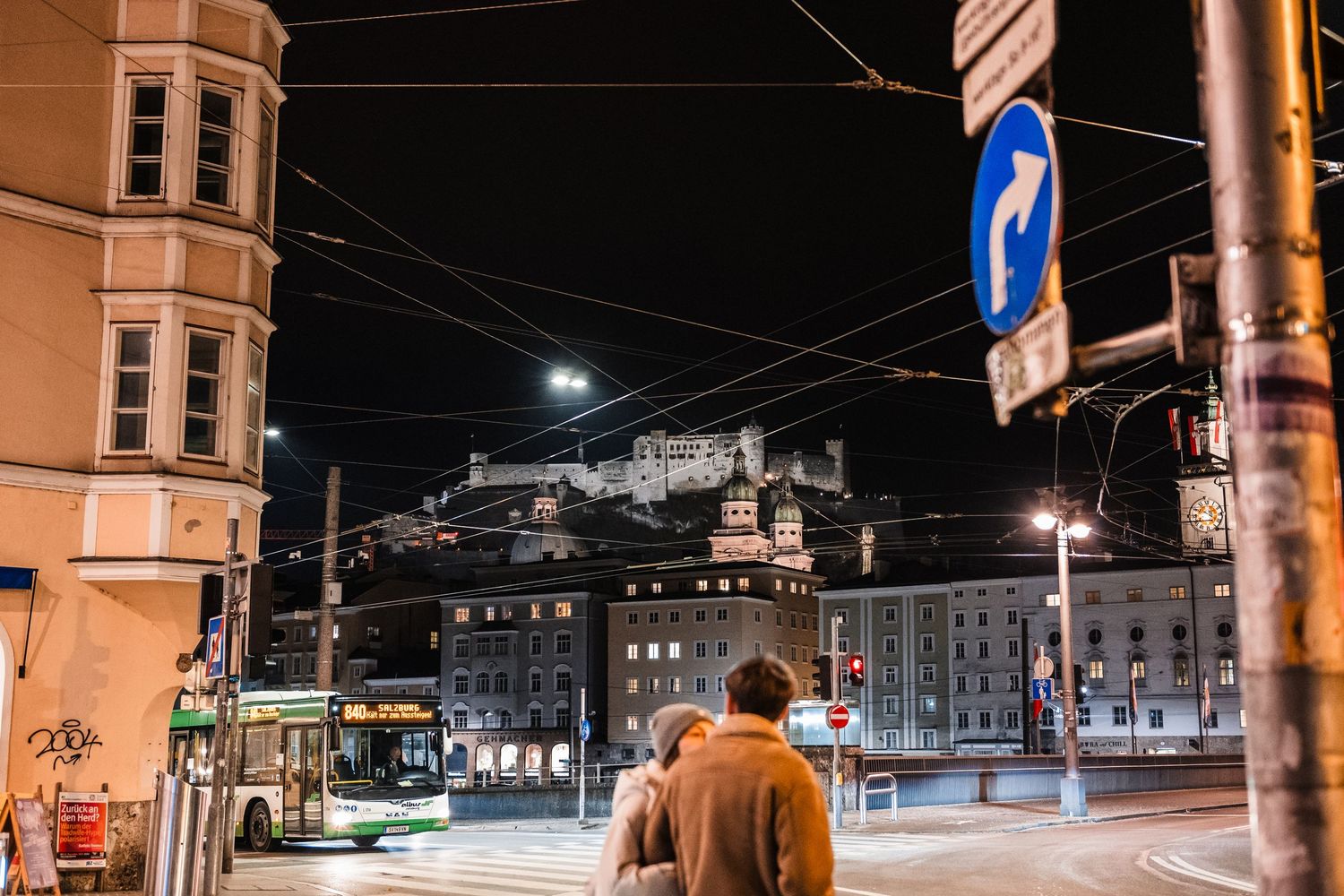 Nighttime urban street scene with illuminated buildings, traffic lights, and utility poles casting warm glows.