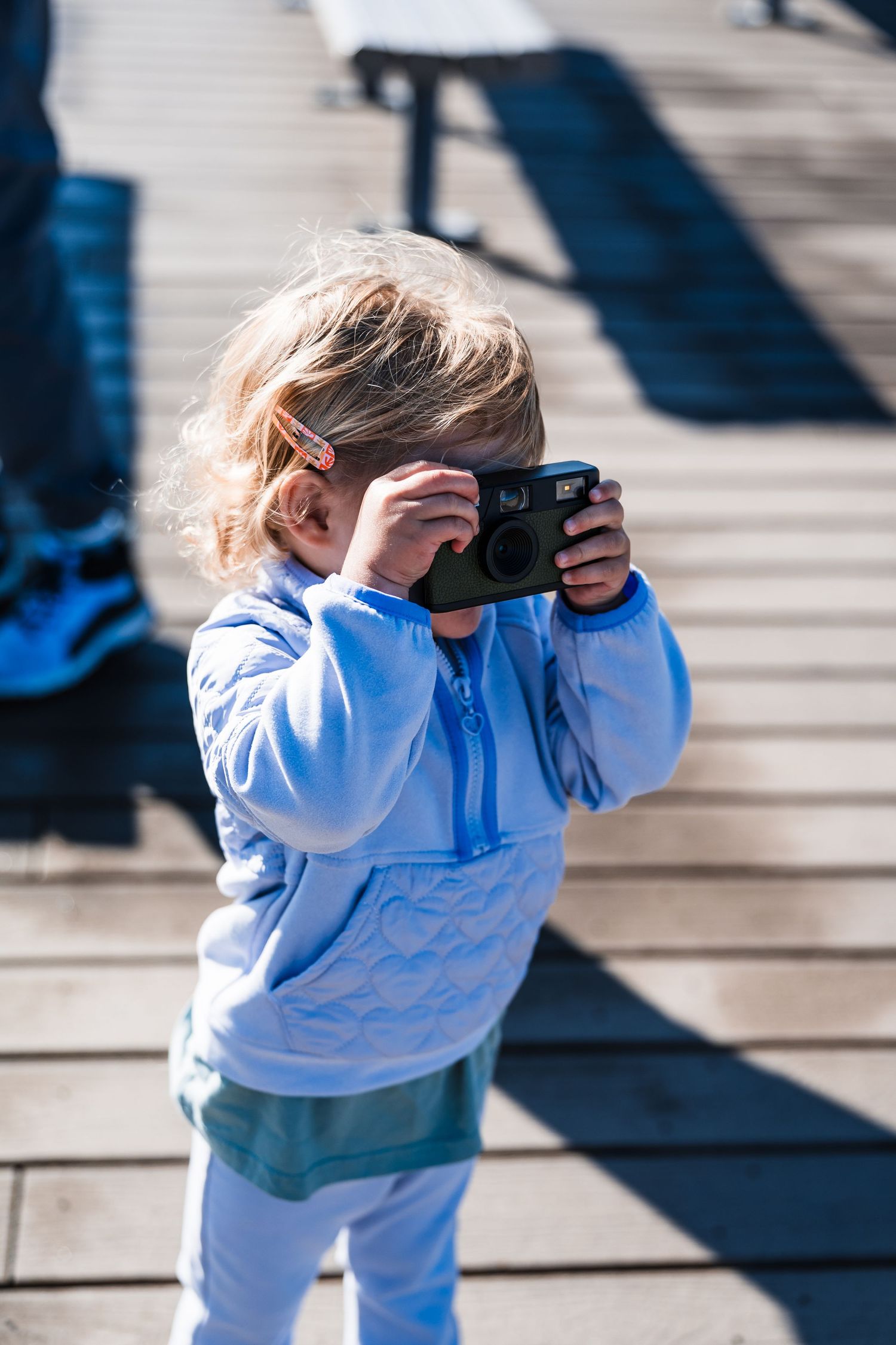 Young child in light blue hoodie and white pants takes a photo with a camera on wooden deck steps.