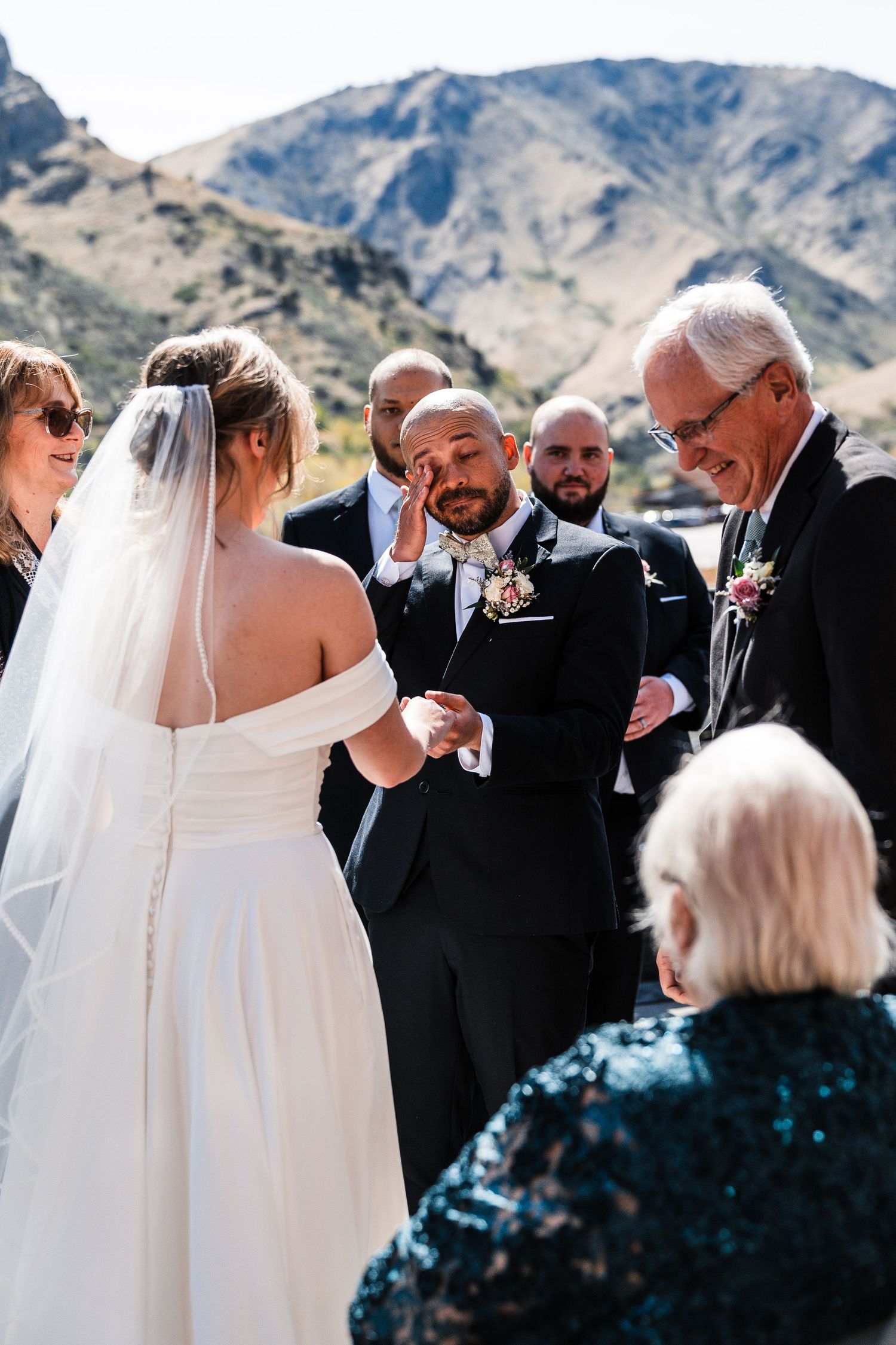 Groom wiping tears during outdoor mountain wedding ceremony while exchanging rings with bride.