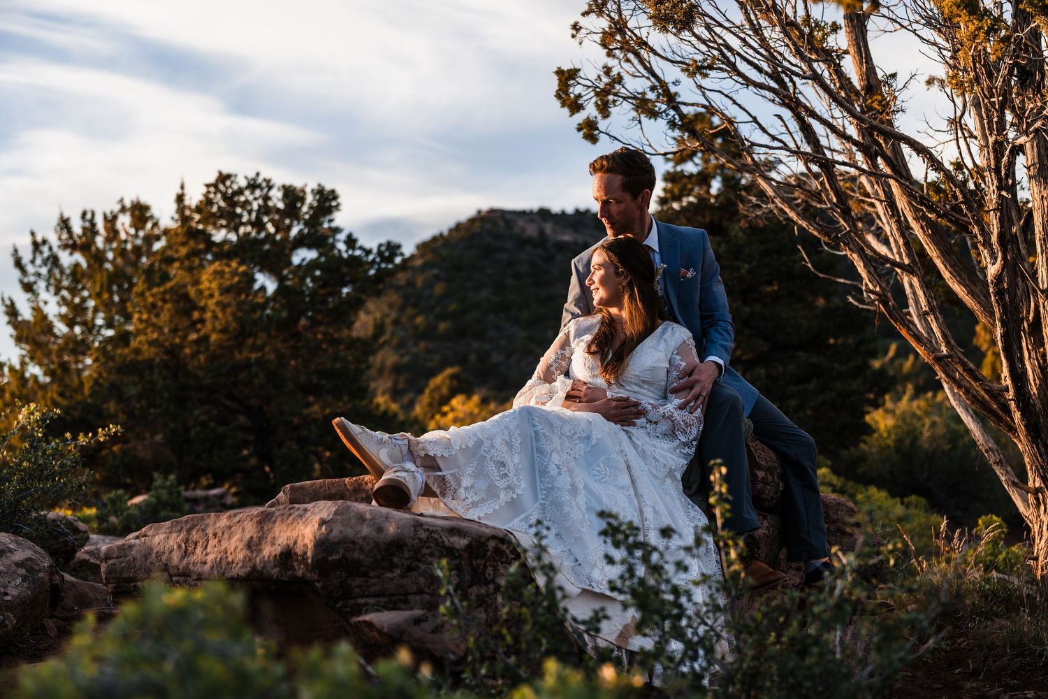 Groom in blue suit and bride in lace gown sitting together on rocks surrounded by golden sunset scenery.