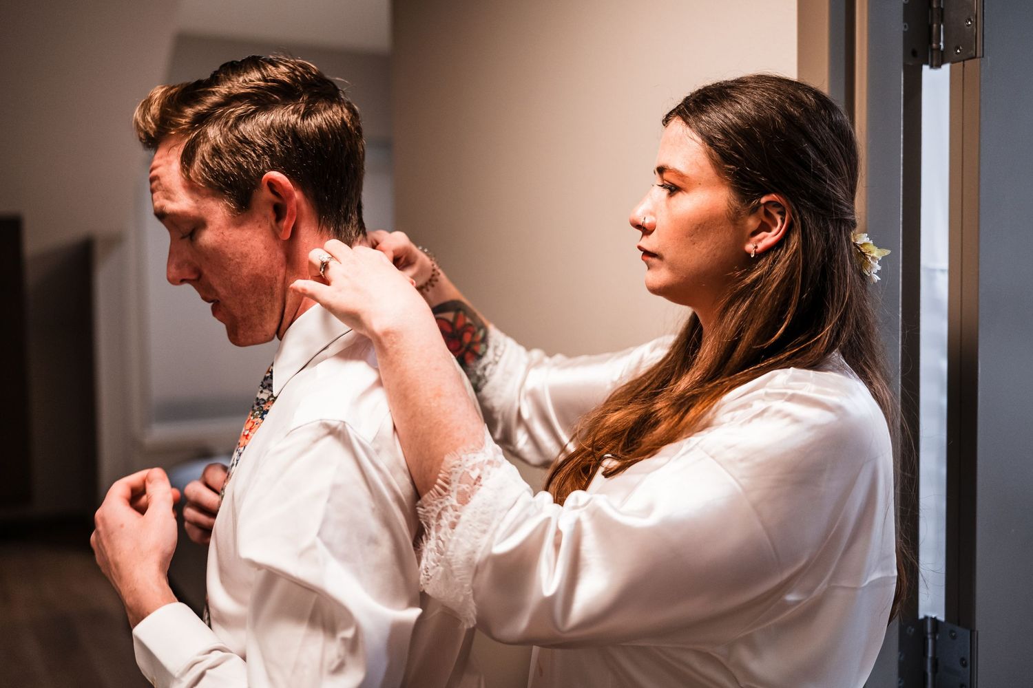 Woman in white robe helping groom button his dress shirt during wedding preparations indoors.