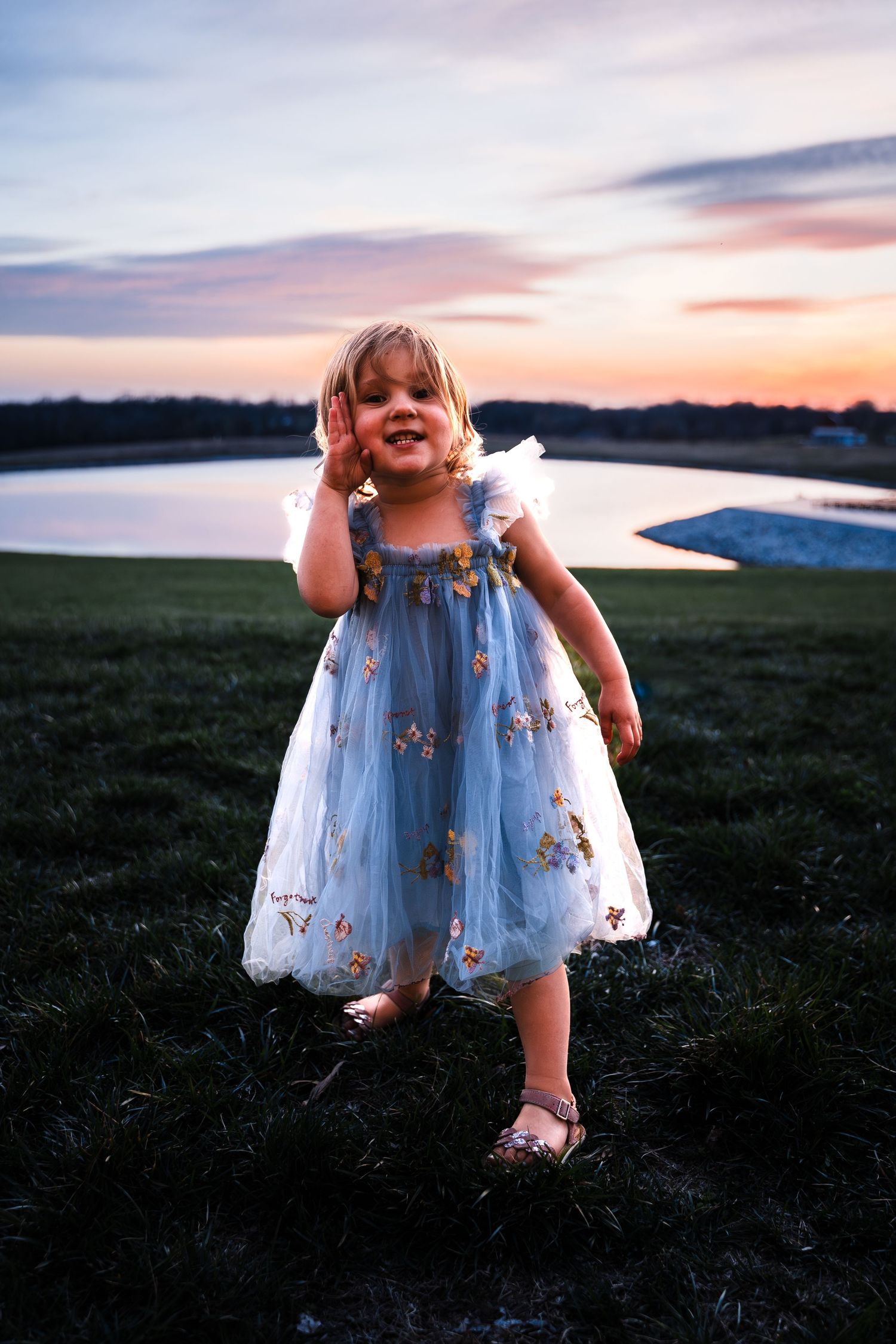 Sweet little girl in blue floral tulle dress standing by pond under vibrant purple sunset sky.