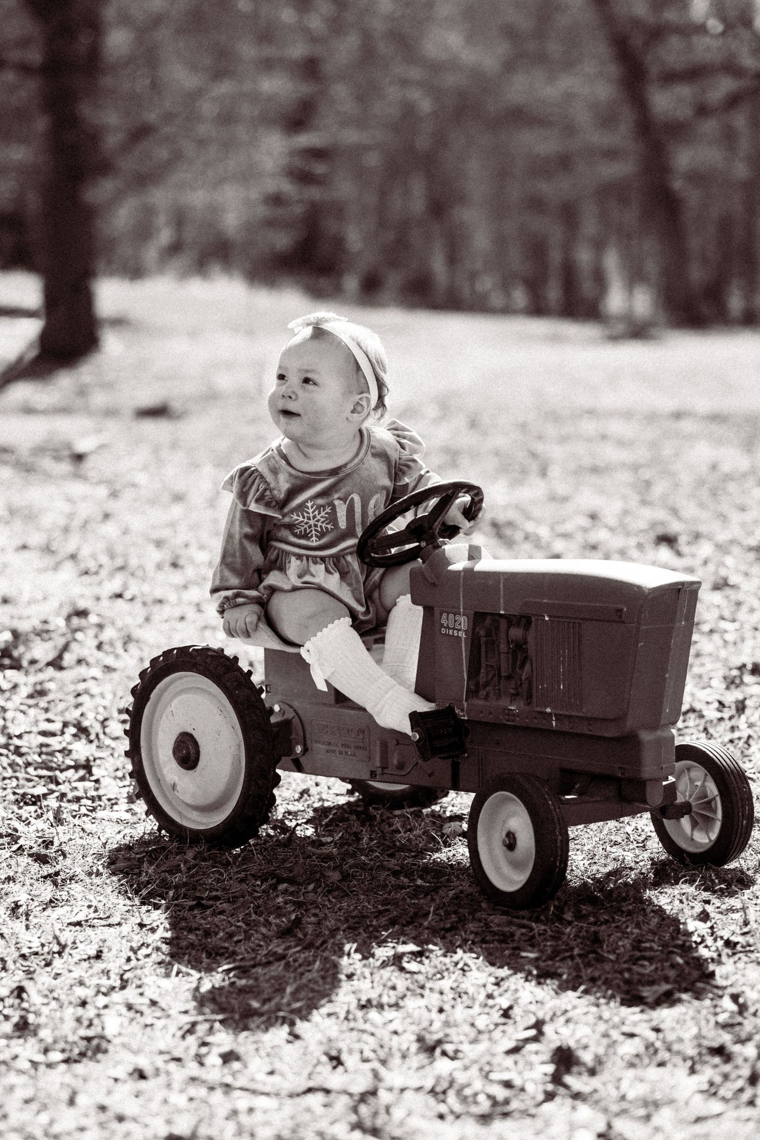 A small child rides a vintage pedal tractor toy outdoors in a black and white photograph.