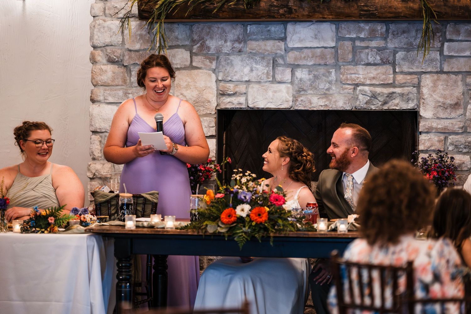 Bridesmaid in lavender dress giving toast speech at wedding reception near stone fireplace.