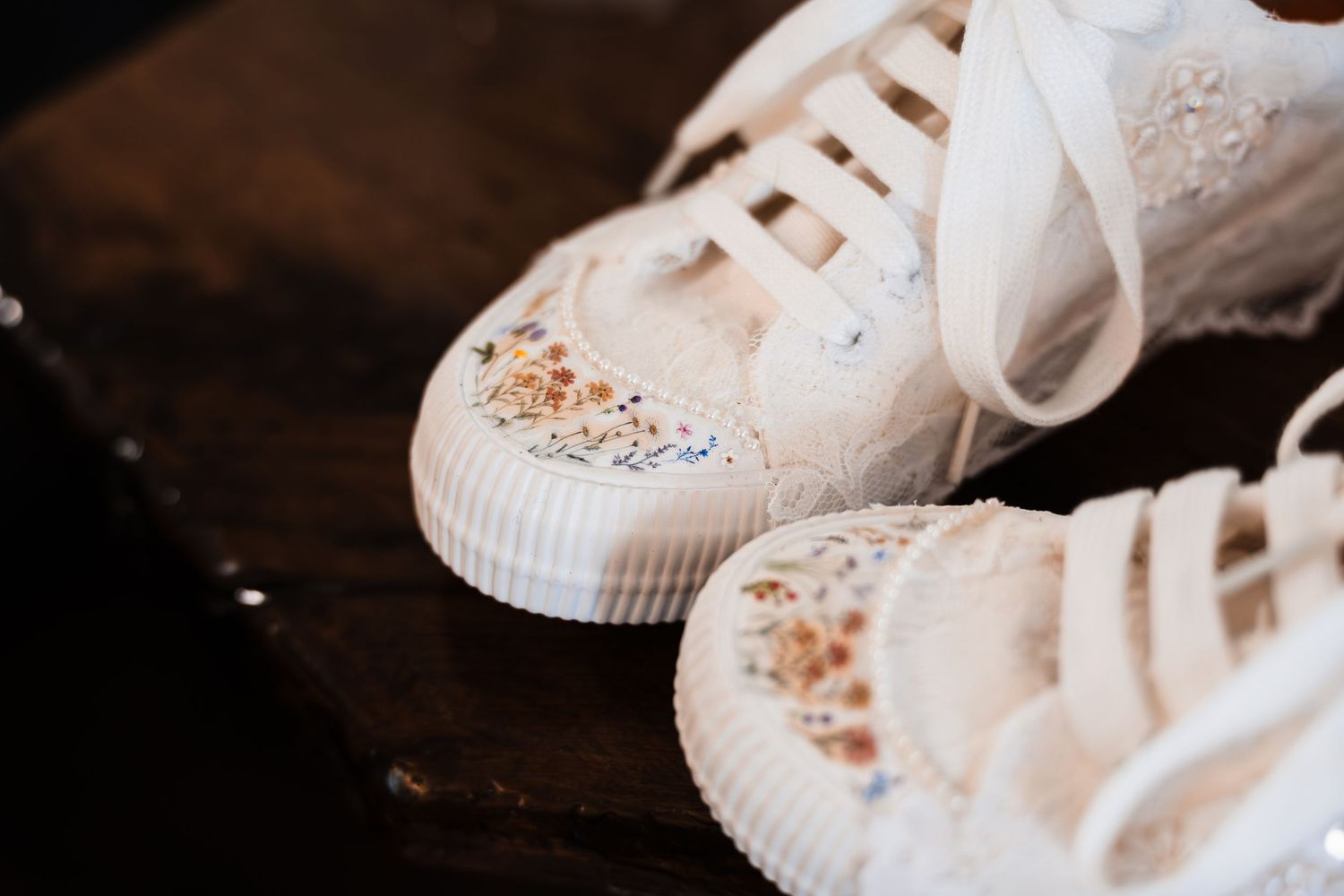 White embellished wedding sneakers with floral details and lace accents displayed on a dark wooden surface.