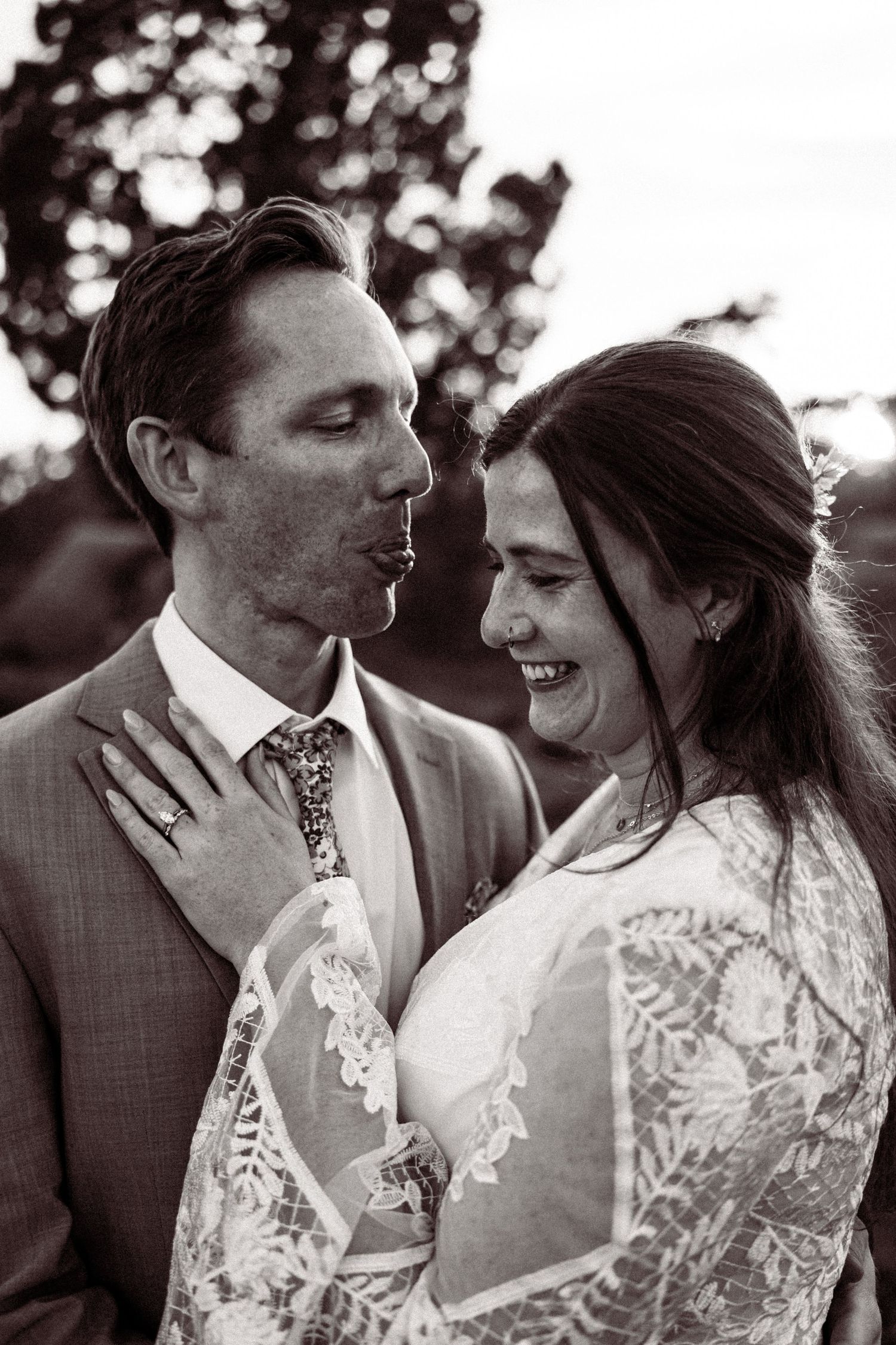 Black and white portrait of groom kissing brides cheek as she laughs, both in wedding attire outdoors.