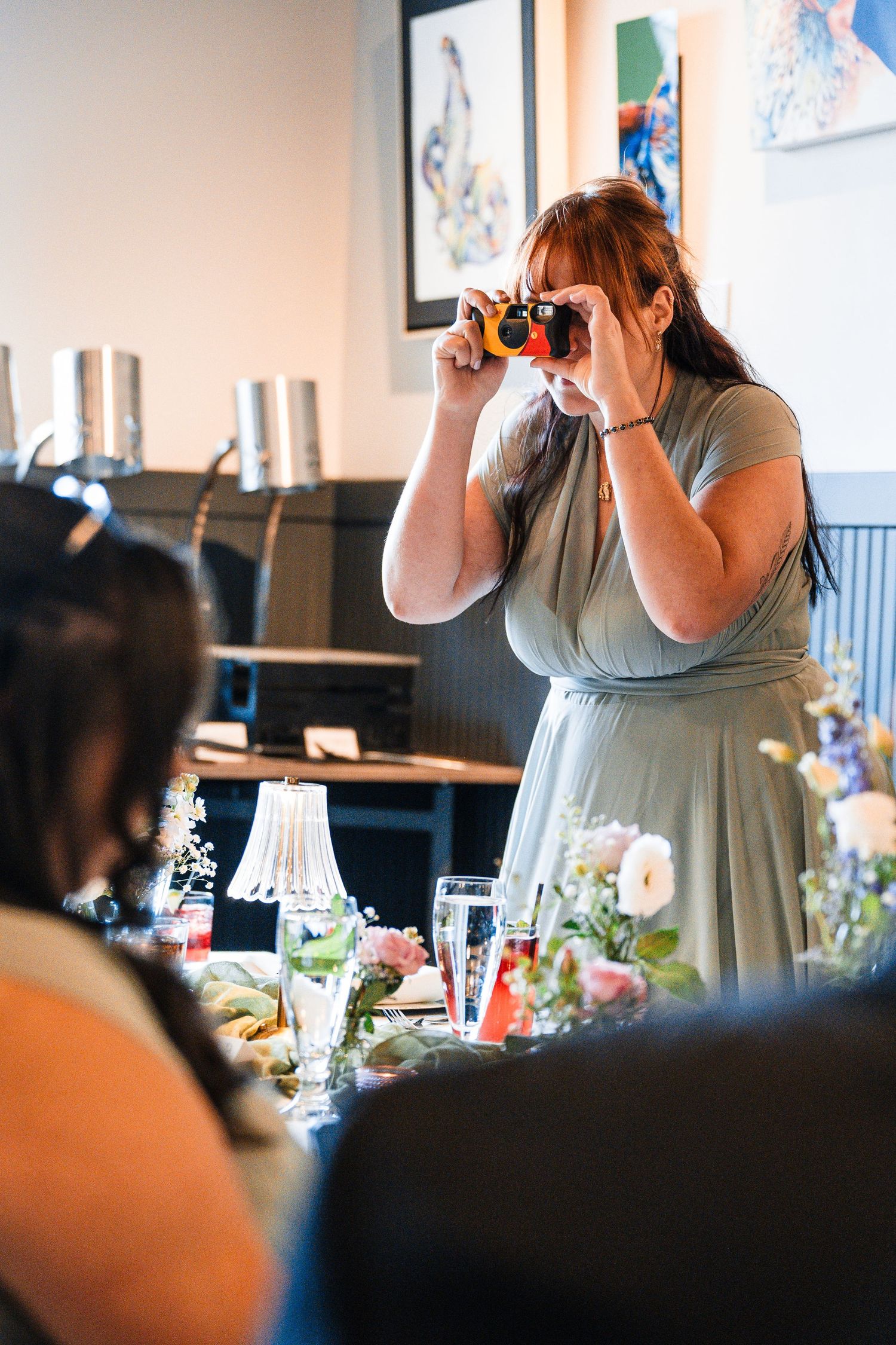 Bridesmaid in sage green dress photographs wedding reception guests with colorful camera.