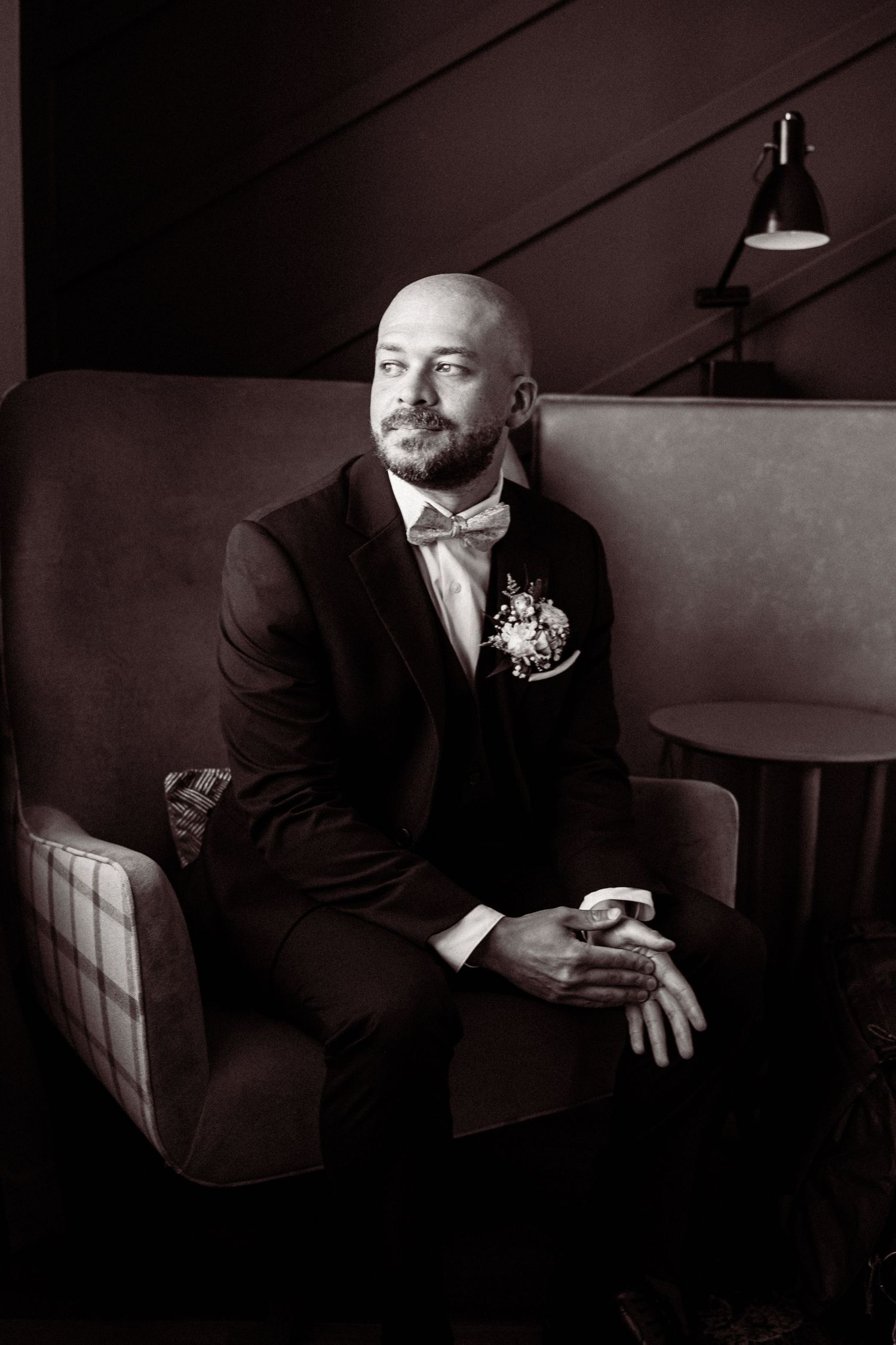 Bald groom in black tuxedo with bow tie and boutonniere sits in armchair, sepia-toned portrait.