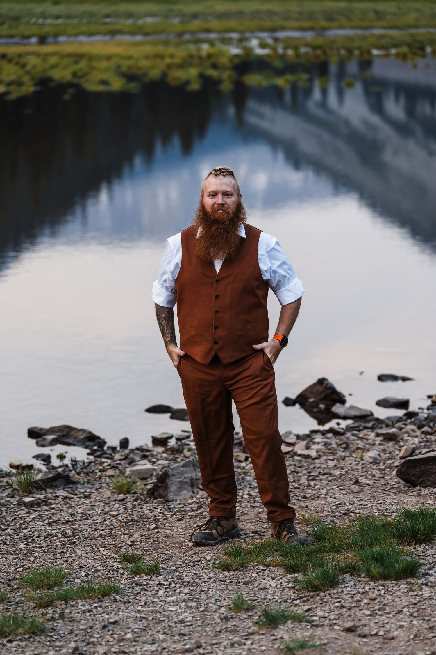 Bearded man in brown vest and trousers stands confidently by a calm reflective river in a scenic outdoor setting.