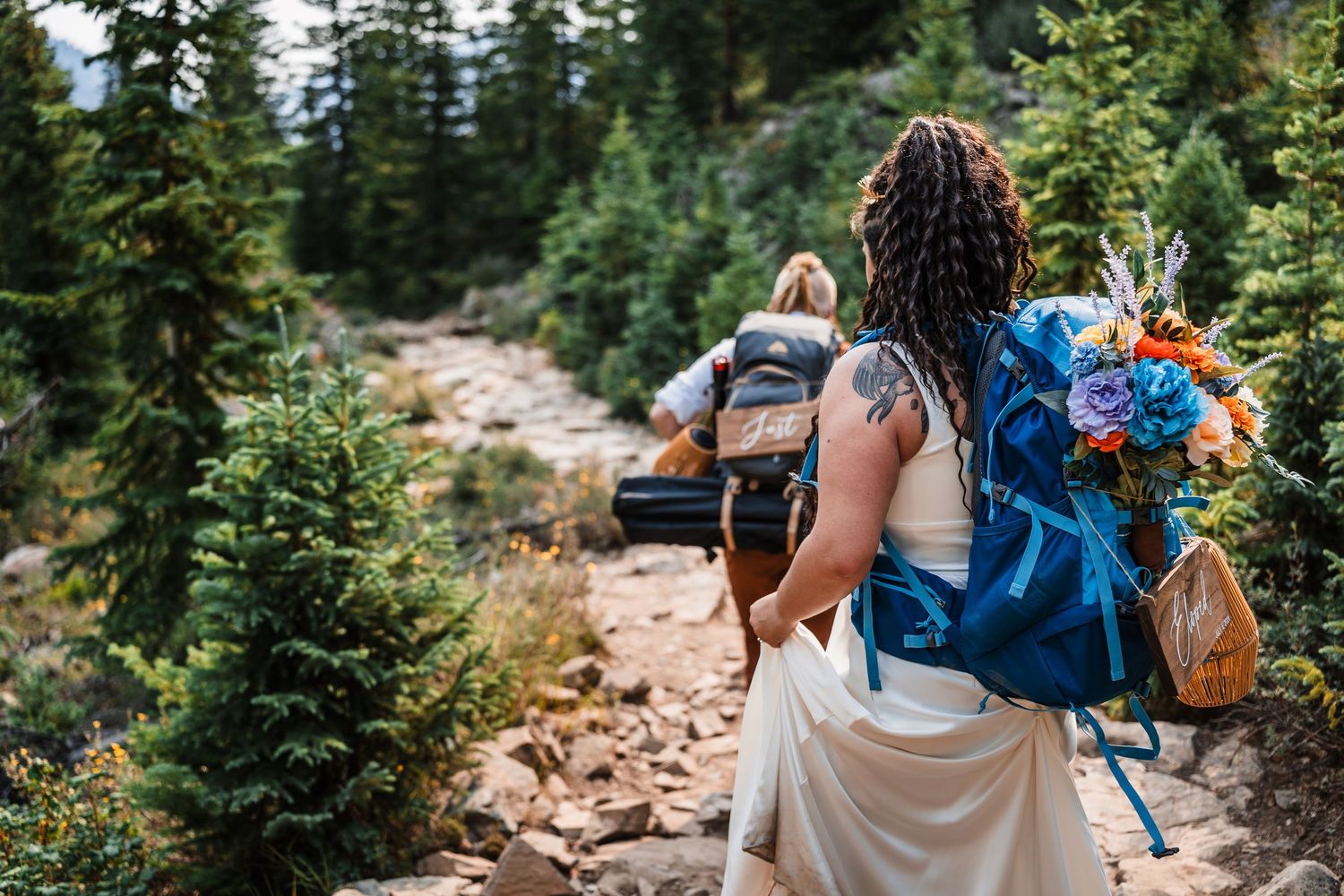 Bride in white dress with blue backpack and colorful bouquet hiking rocky mountain trail through pine forest.
