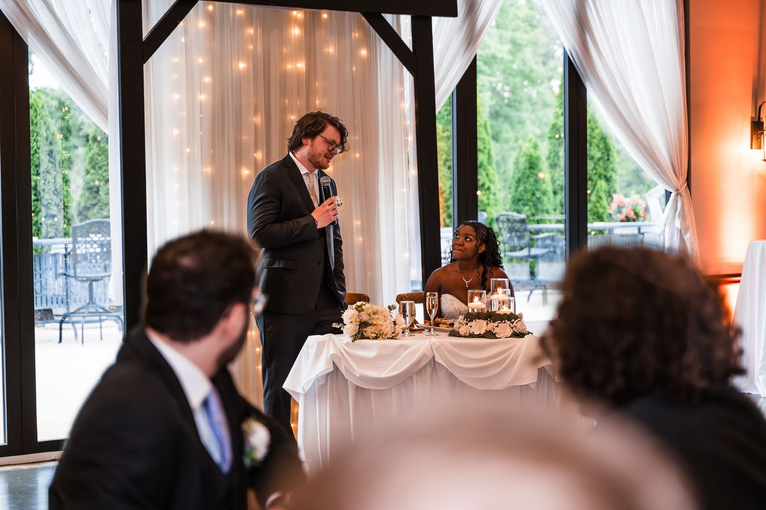 Groom giving a heartfelt speech at wedding reception while bride listens at the sweetheart table.