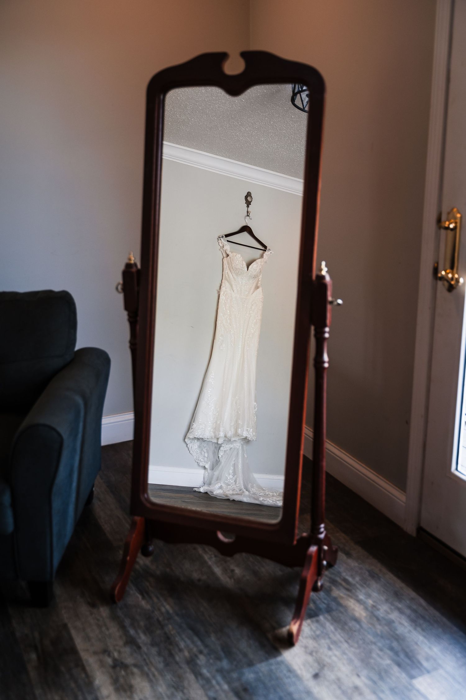 White wedding dress reflected in an ornate wooden cheval mirror on hardwood floors in an elegant room.