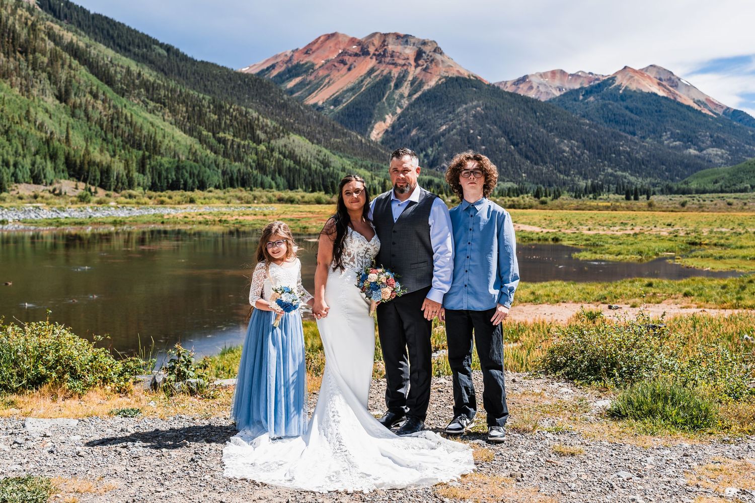 Wedding family portrait with bride, groom, and two children posing before a stunning red mountain and alpine lake.