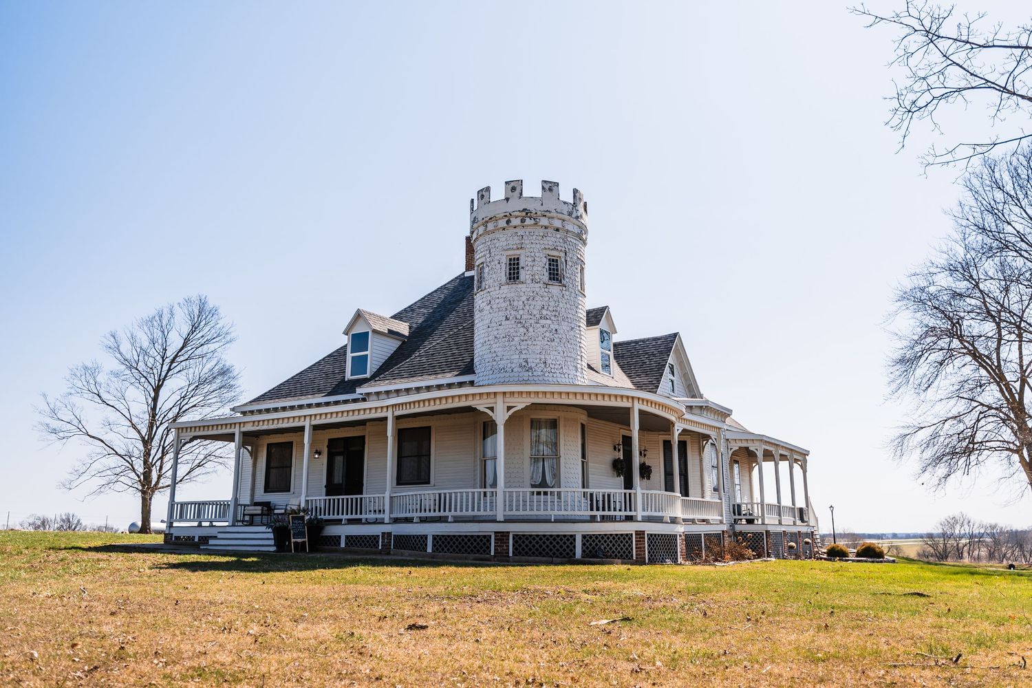 Historic castle-style Victorian home with wrap-around porch and stone turret on a grassy hill under clear blue sky.