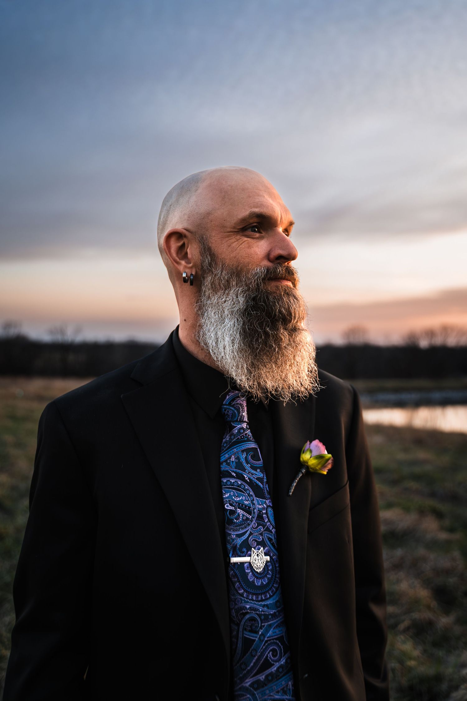 Bald man with long gray beard wearing black suit and floral boutonniere stands in field at sunset.