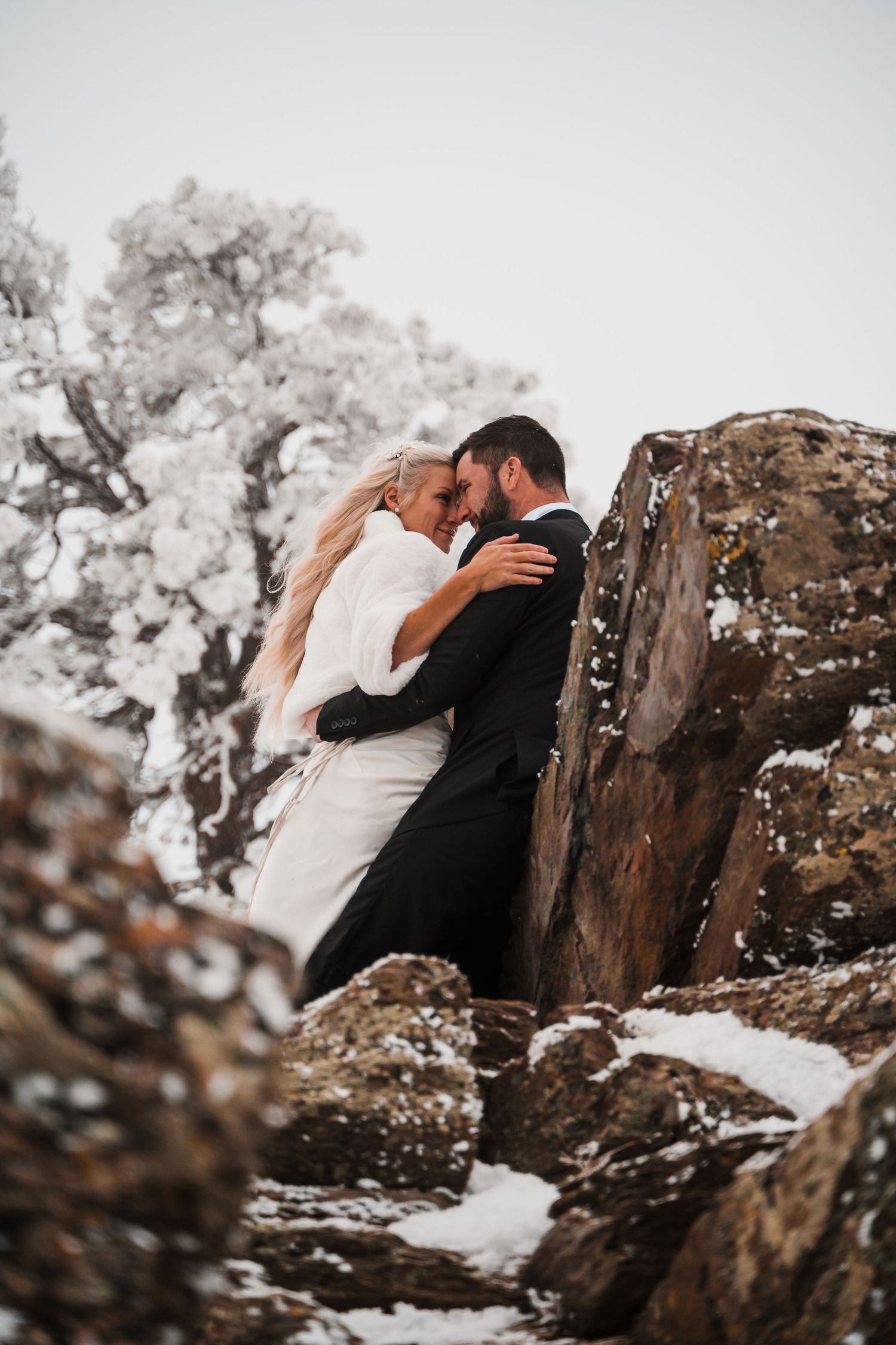 A romantic wedding moment captured against rocky cliffs in winter, with the couple sharing an intimate embrace.