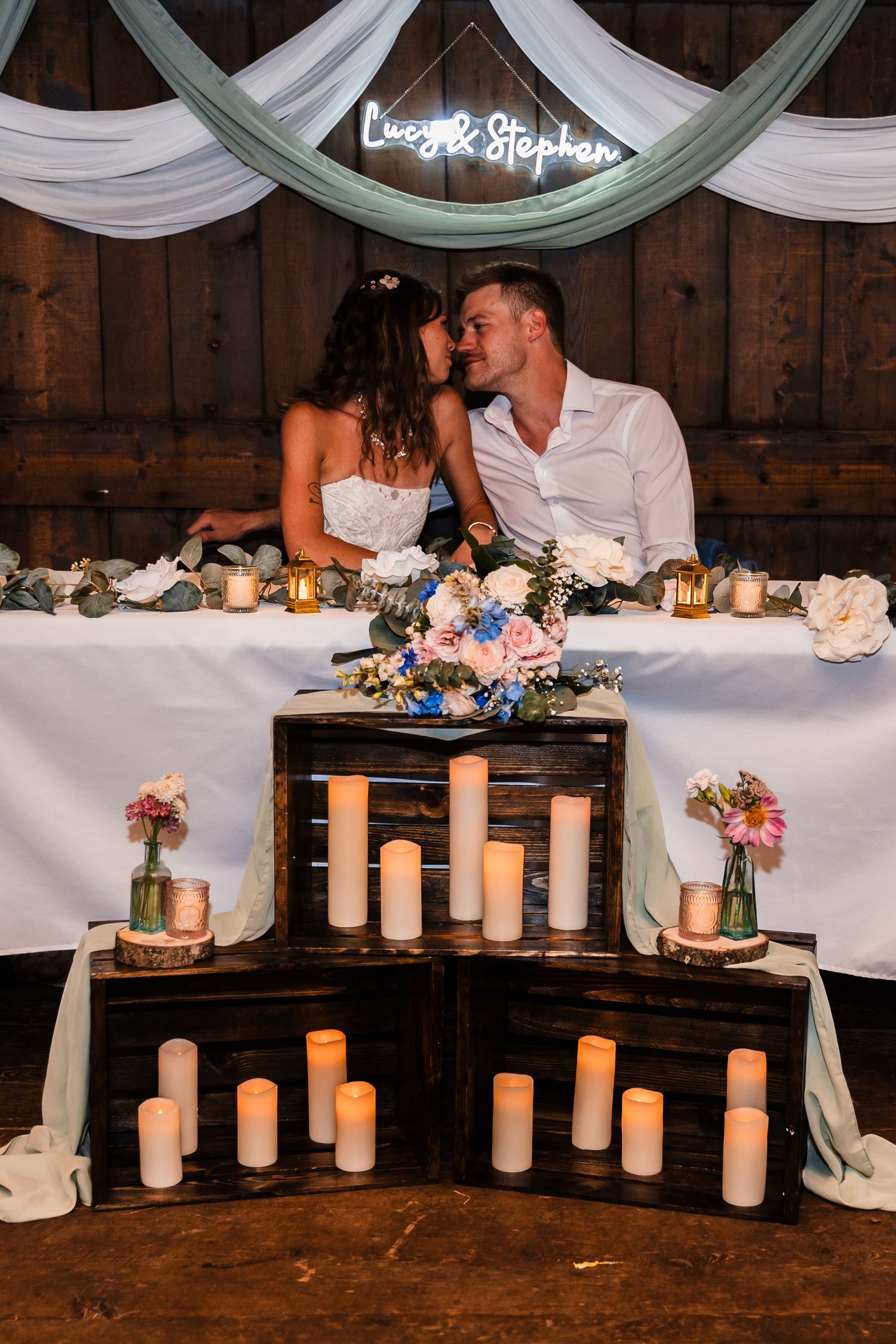 Romantic reception setup with tiered candles and floral decorations under draped fabric and neon sign.