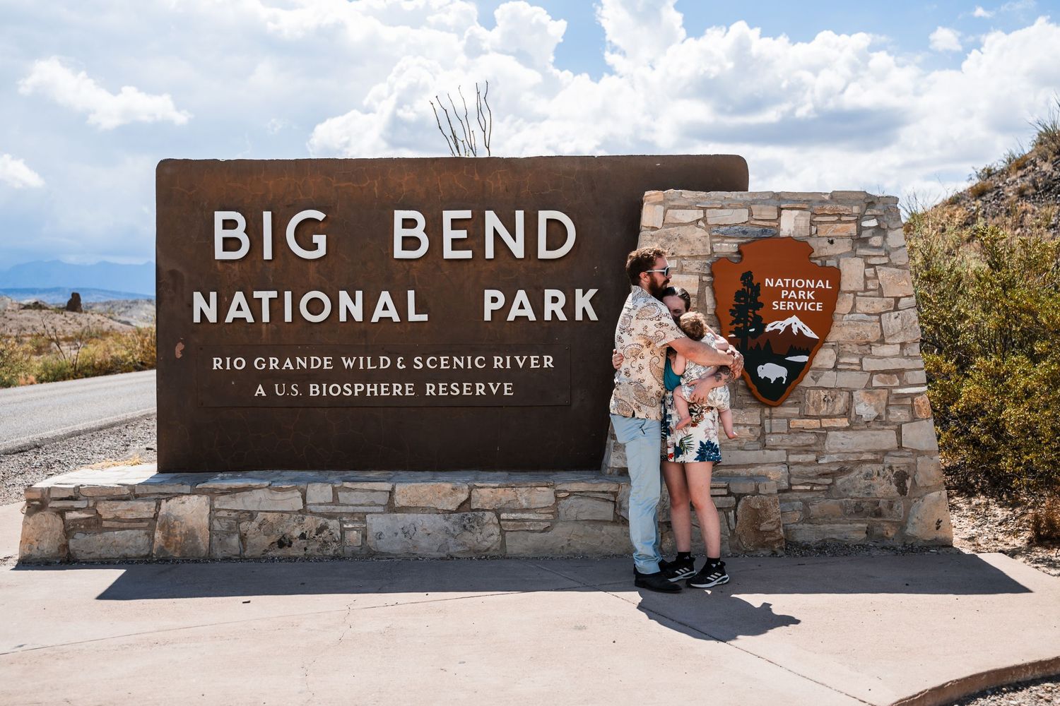 Visitors stand at the entrance sign of Big Bend National Park in Texas against a backdrop of mountains and blue sky.