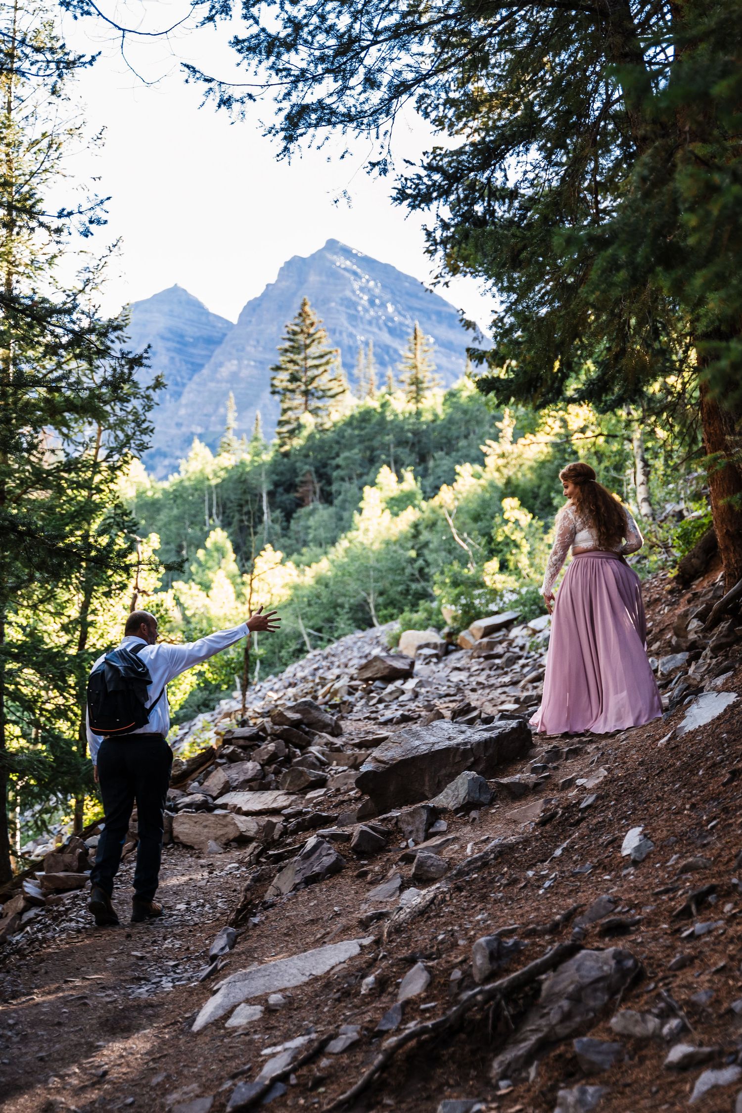 Pink-dressed figure poses on rocky mountain trail while being photographed.