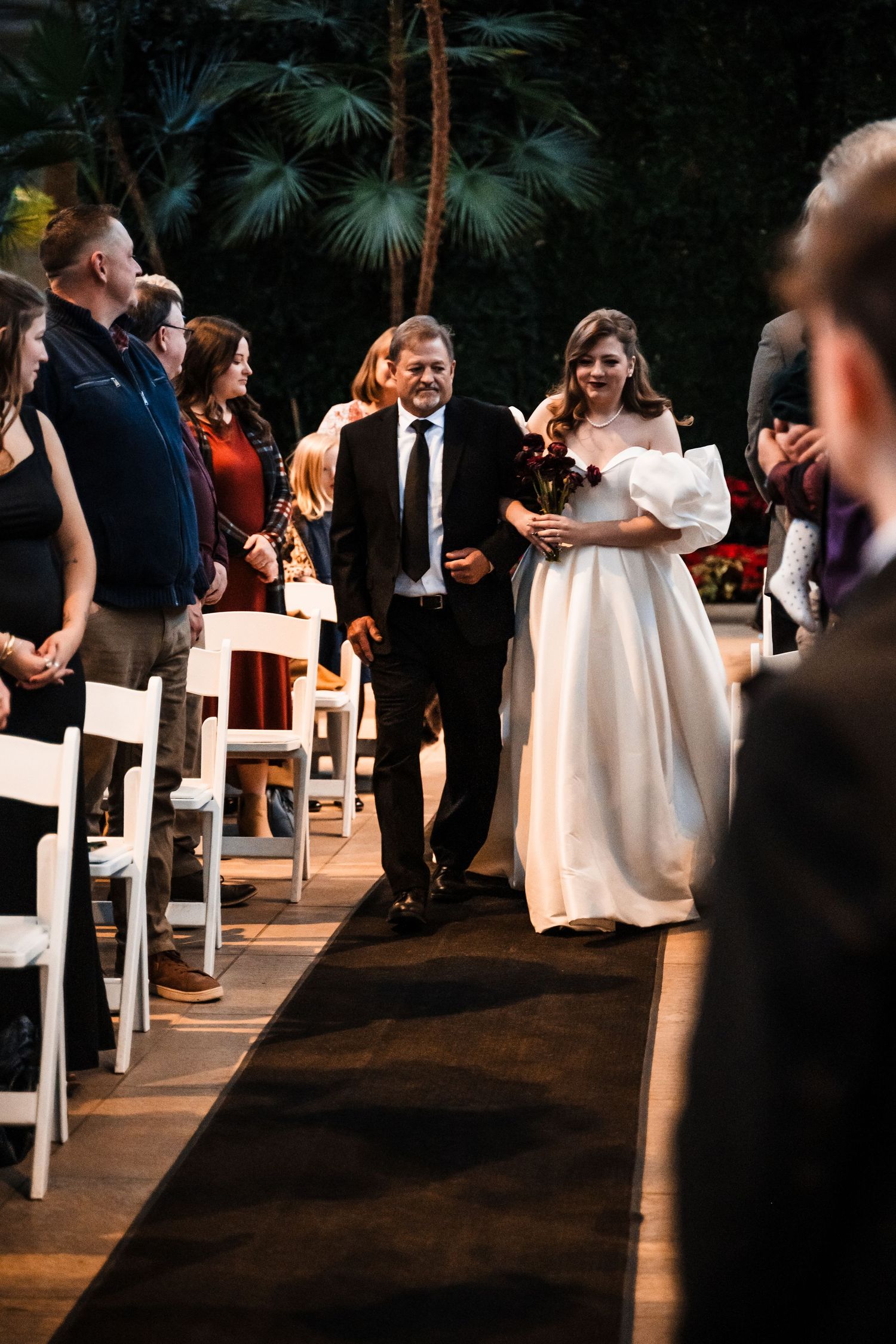 Wedding ceremony processional in an indoor venue with palm trees and white chairs lining the aisle.