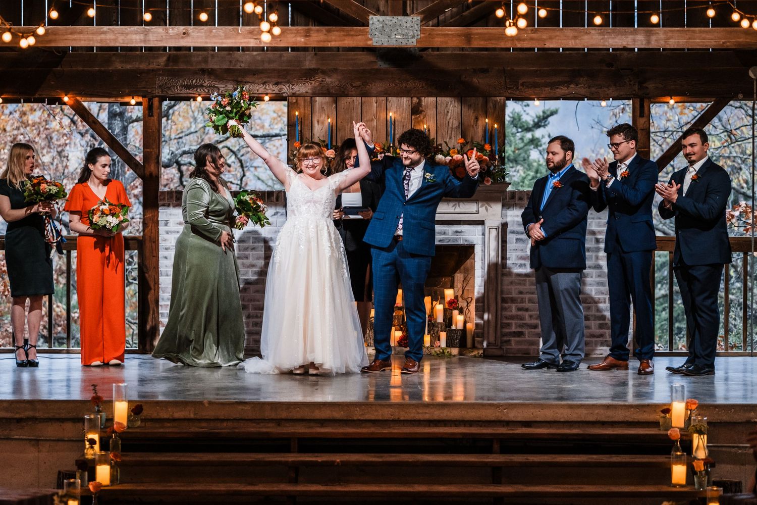 Wedding celebration with bridal party on elevated platform in rustic venue with string lights.