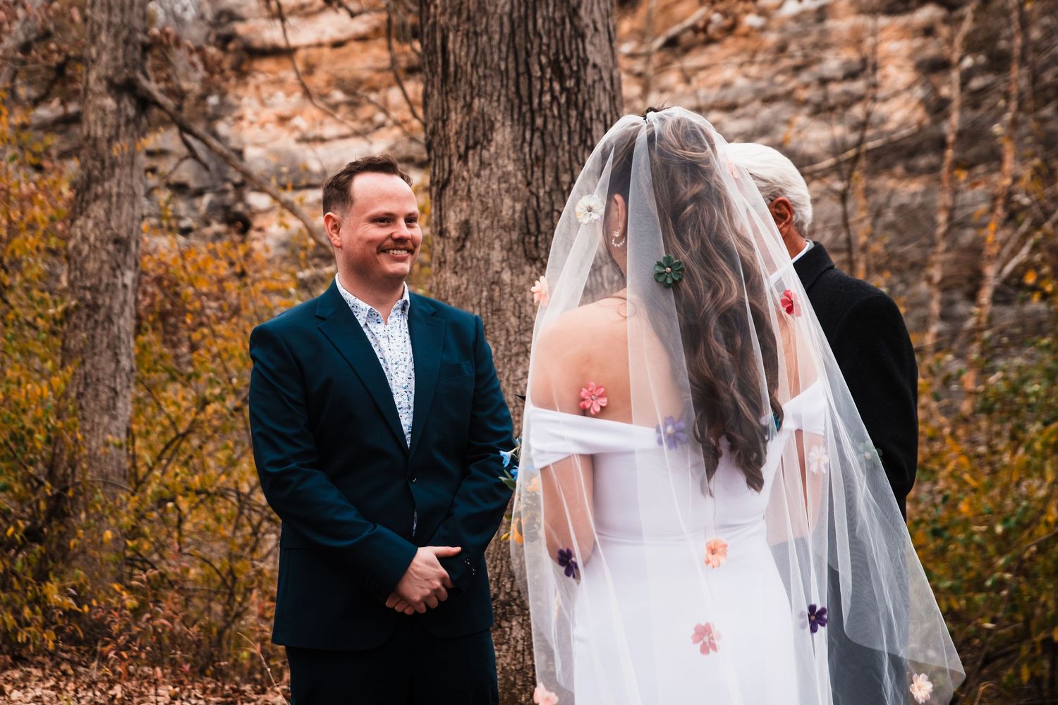A smiling couple exchange vows during an autumn outdoor wedding ceremony with rocky cliffs in the background.