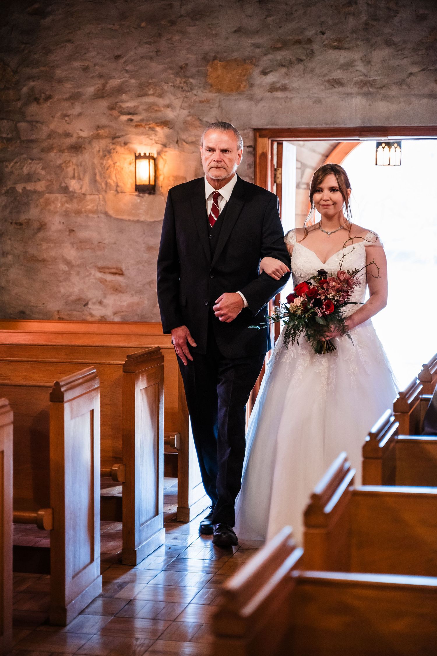 Bride holding red bouquet walks down church aisle in traditional wedding ceremony.