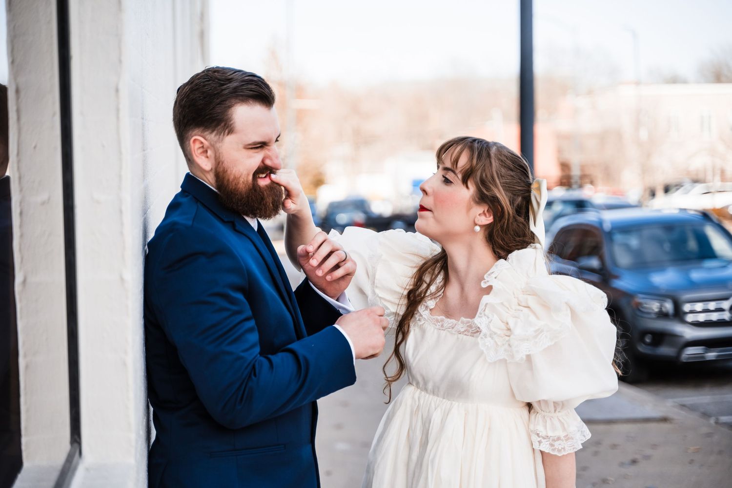 A couple shares a playful moment outdoors, with the person in navy suit and white dress laughing together.