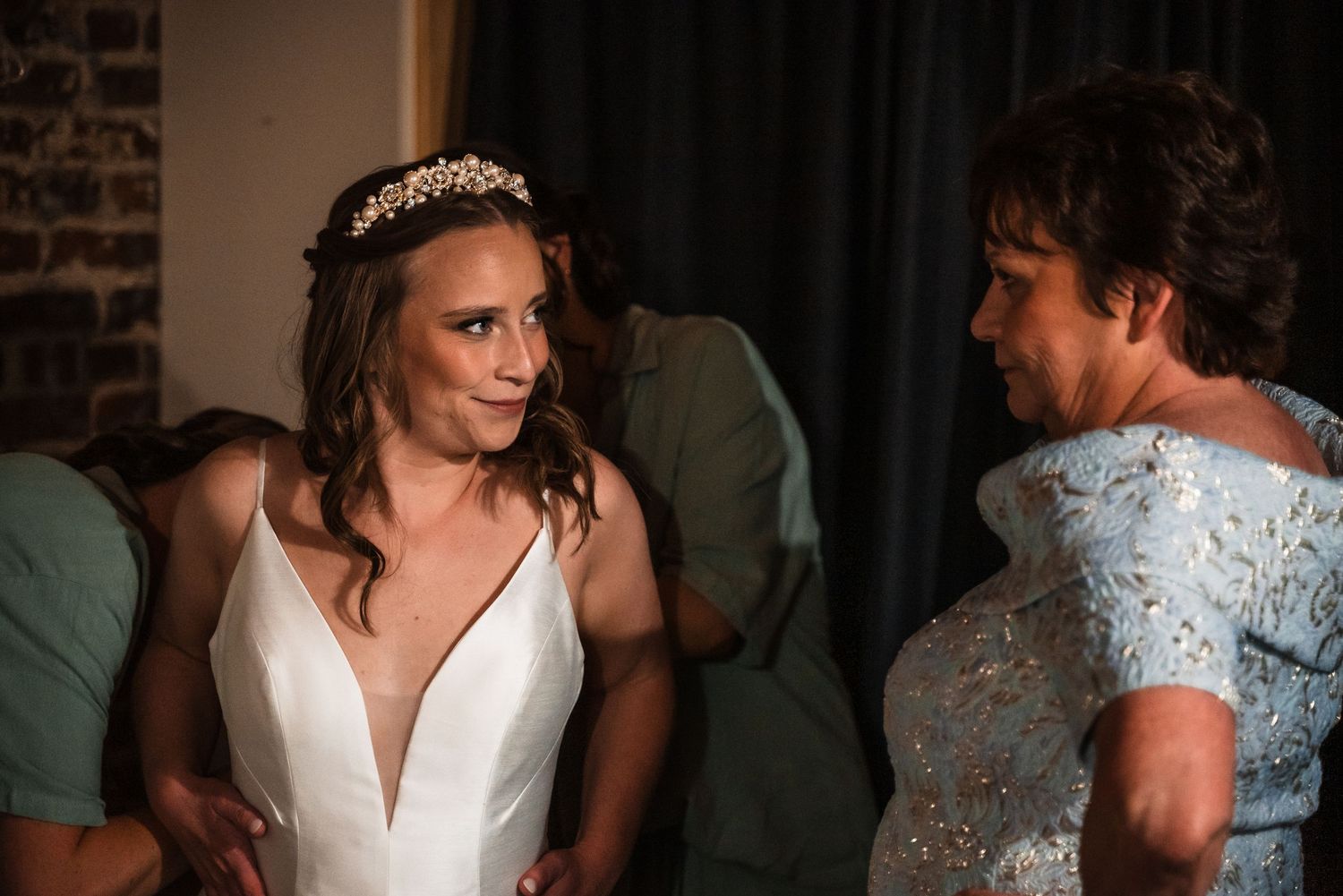 A bride in a white plunging neckline gown wearing a tiara shares a moment with someone in a light blue dress.