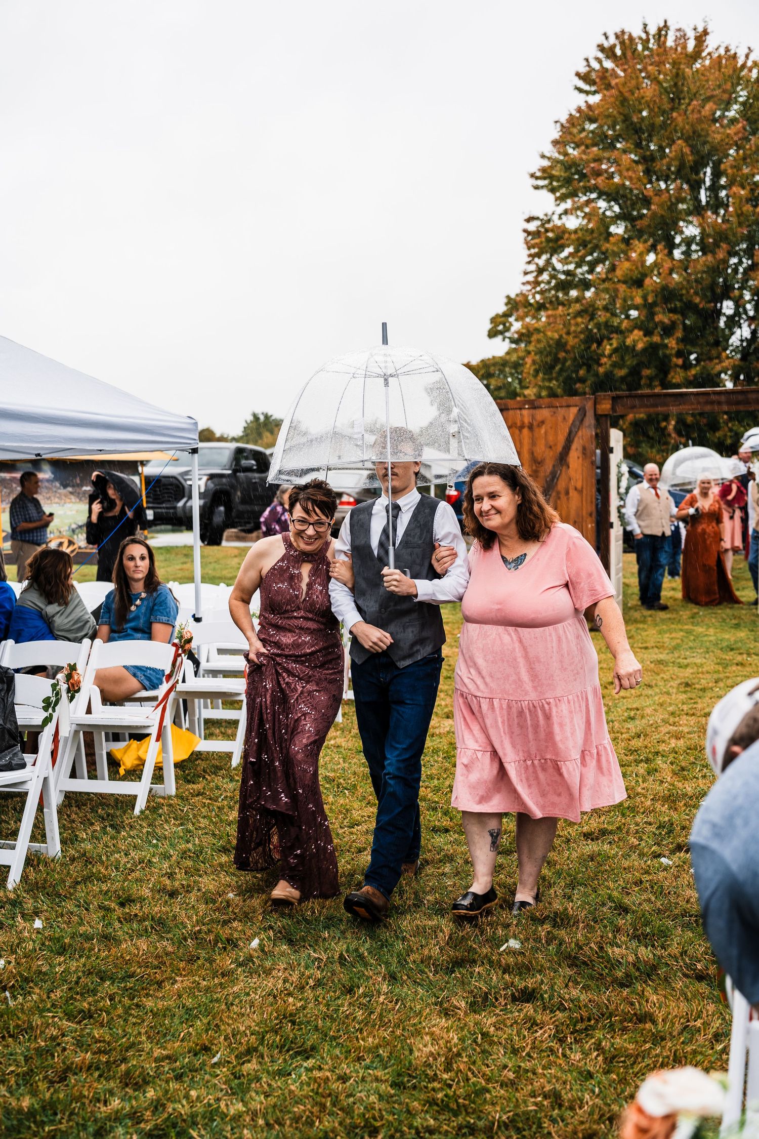 Wedding guests dance and celebrate on a grassy lawn with autumn trees and white tents in the background.