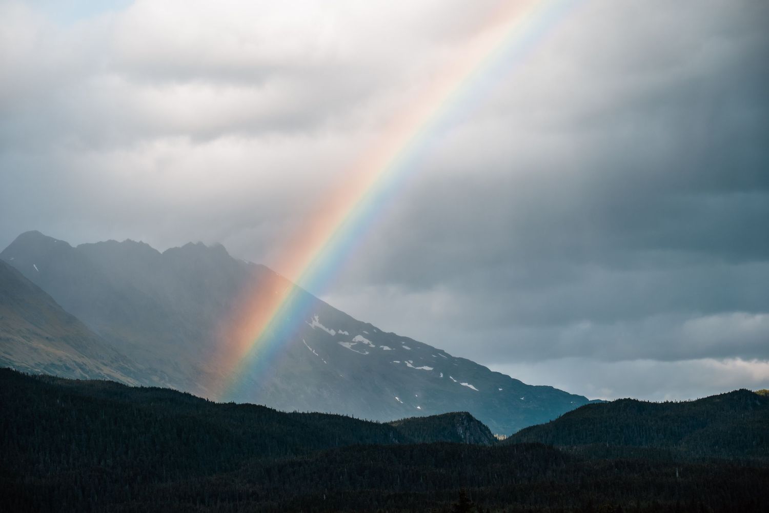 A vibrant rainbow arches over misty mountain peaks against a moody clouded sky.