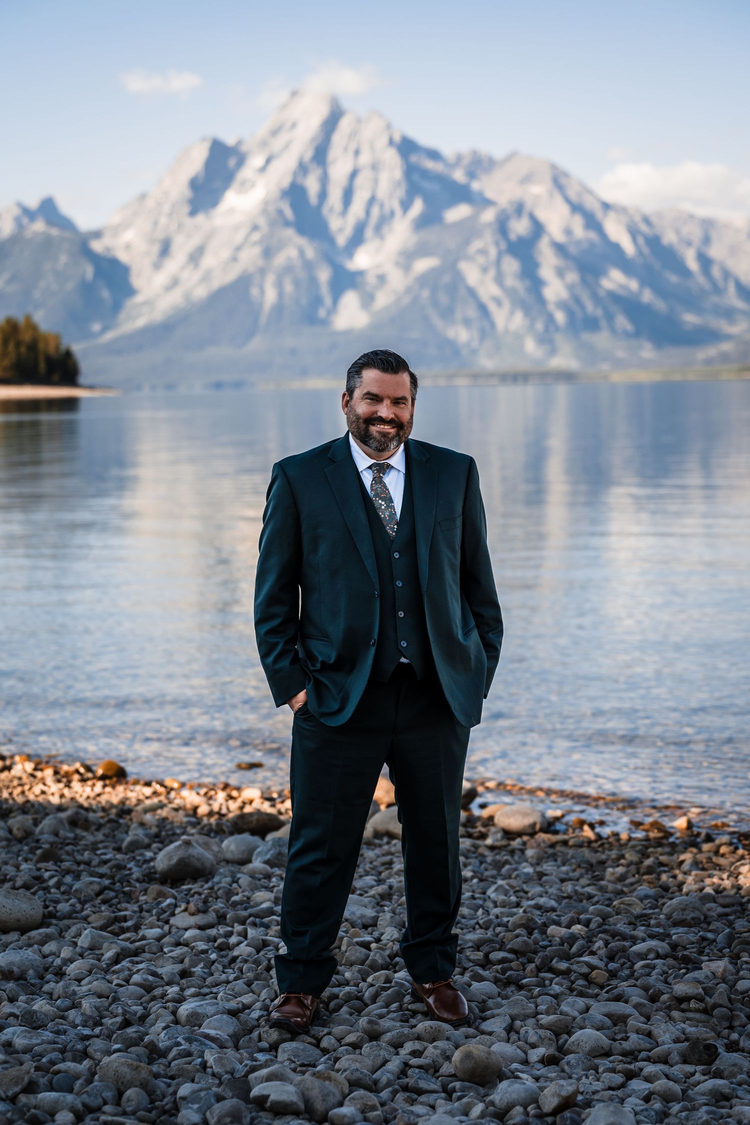 A suited figure stands on a rocky shoreline with snow-capped mountains and a serene lake in the background.