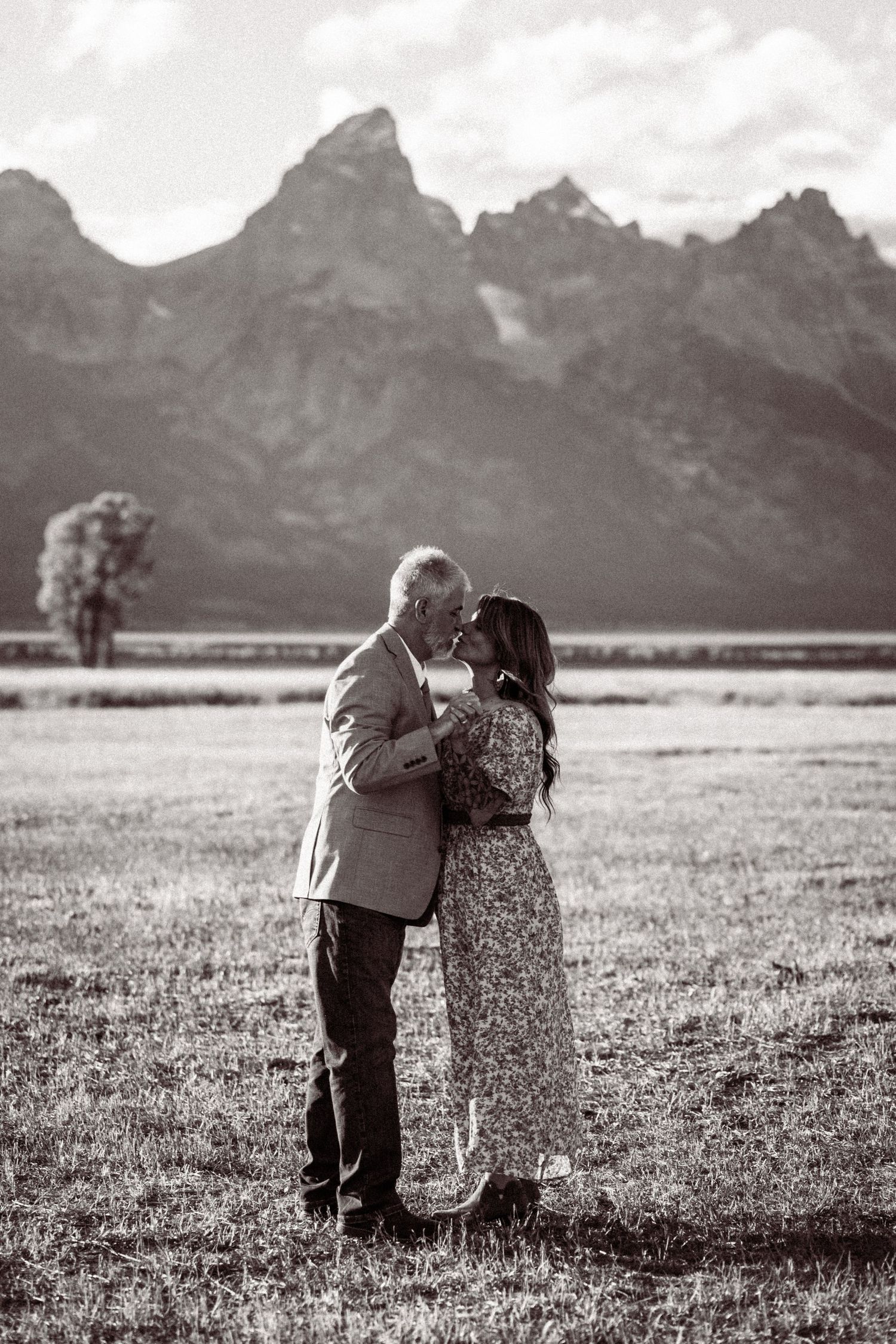Two people share a tender moment in a meadow with majestic mountains in the background.
