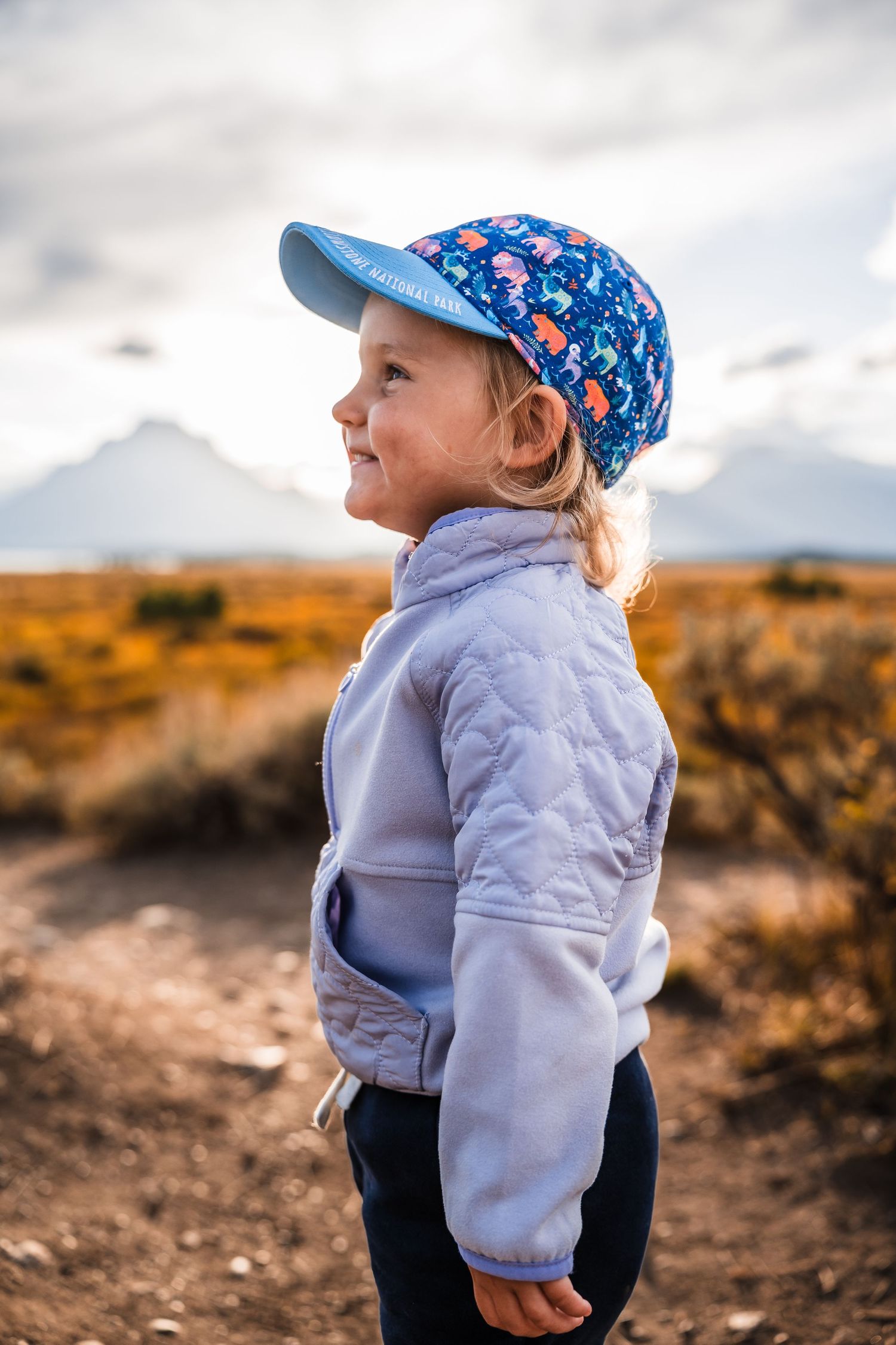 Child in light jacket and blue patterned hat exploring outdoors against cloudy sky.