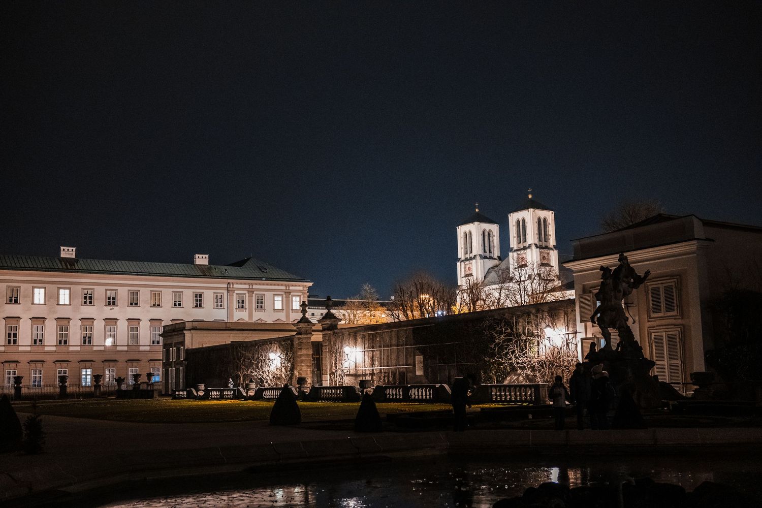 Historic church with twin towers illuminated at night alongside classical buildings reflected in calm water.