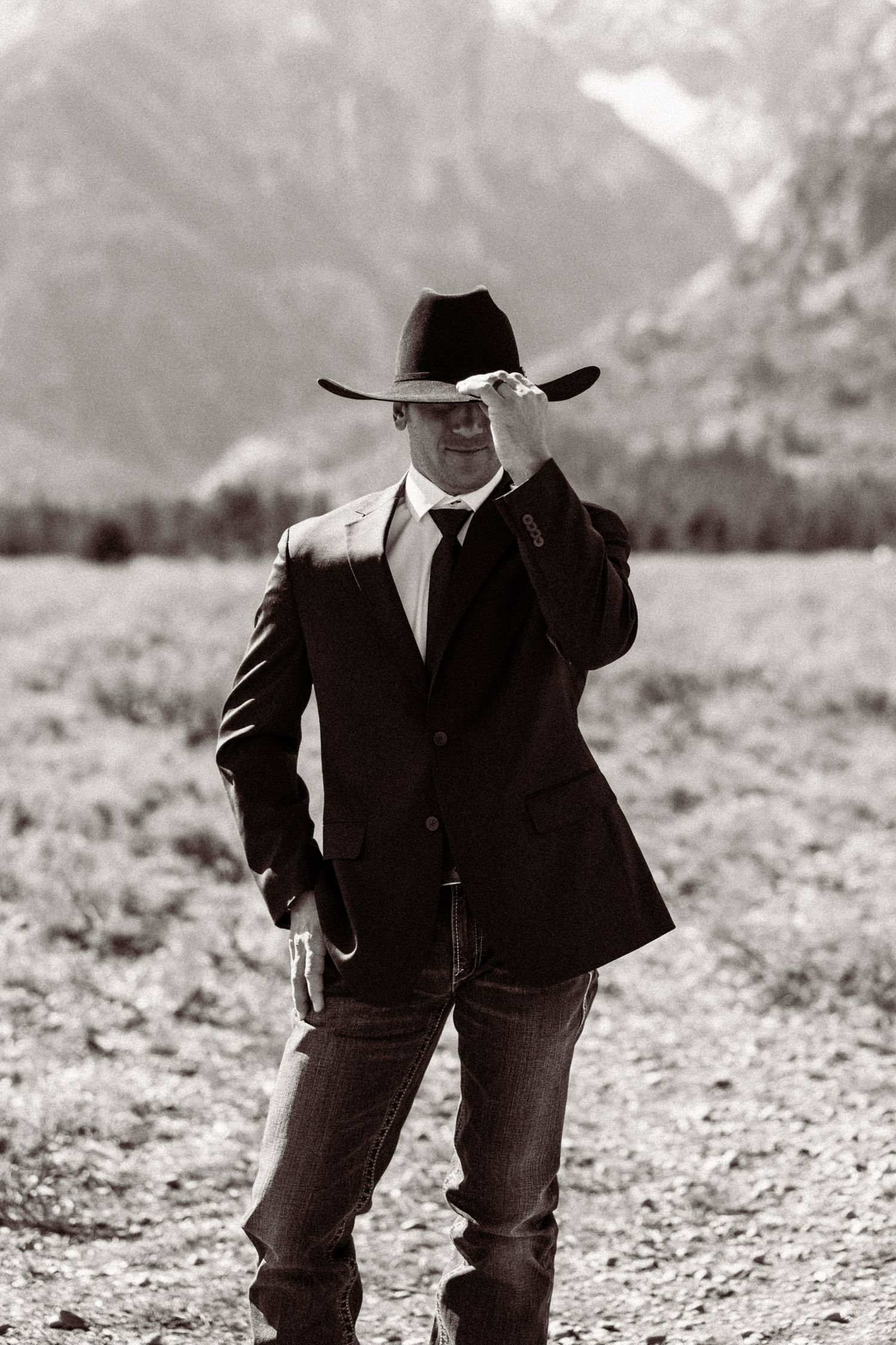 Black and white portrait of a cowboy in a suit and hat standing in a mountain landscape.