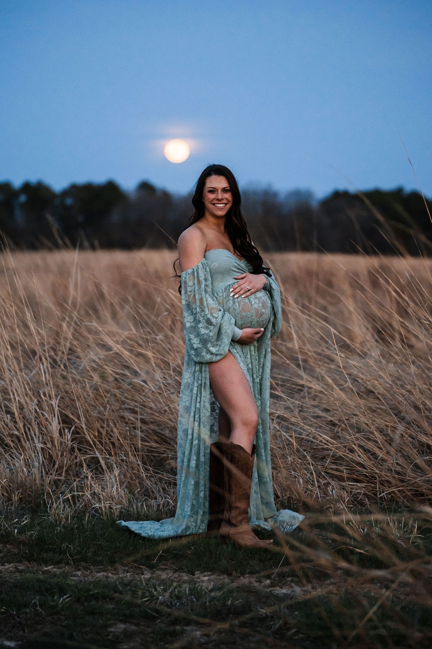 A figure in a turquoise robe poses in tall grass at dusk with a full moon rising in the background.