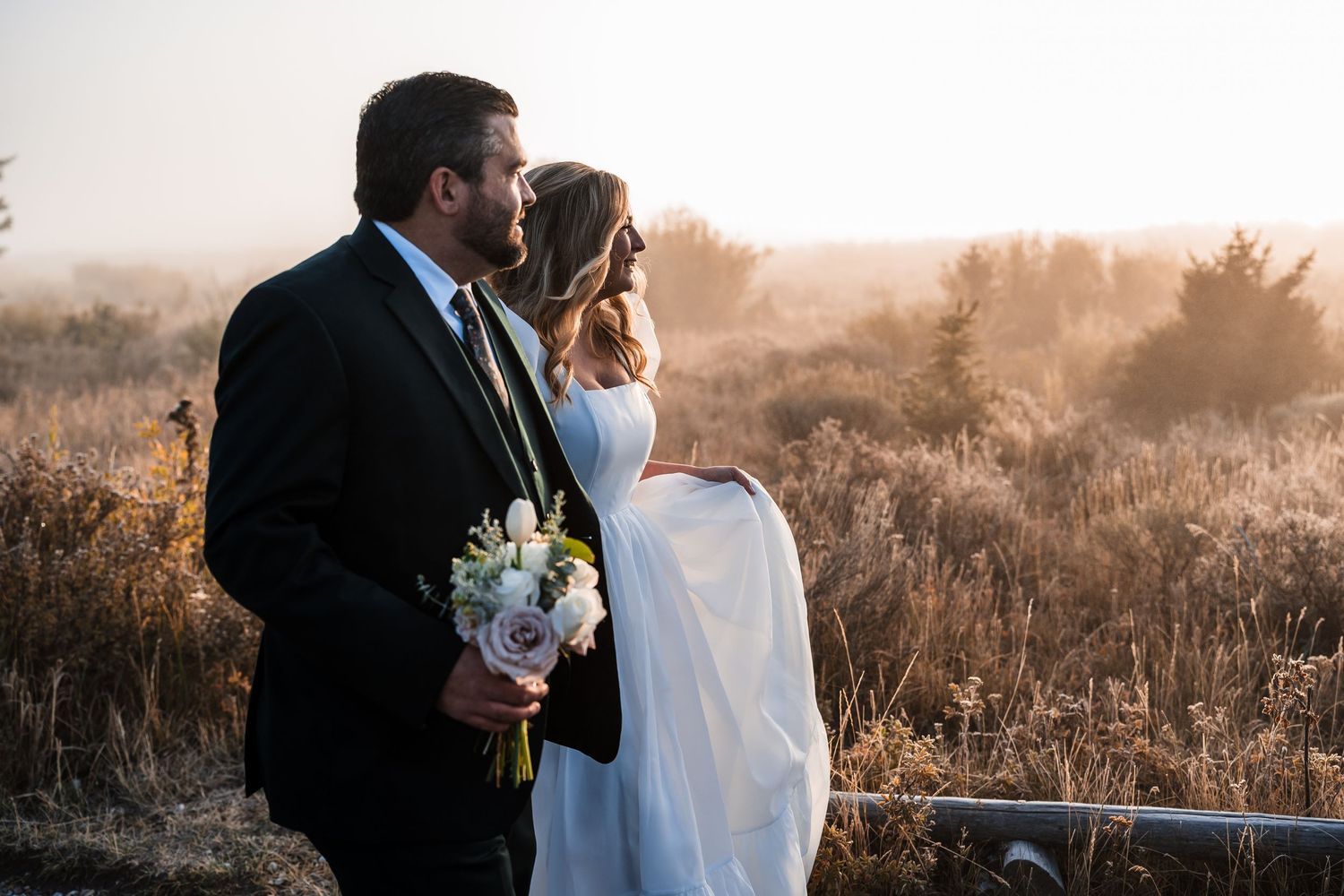 Newlyweds share an intimate moment in a golden field during sunset with mountains in the background.