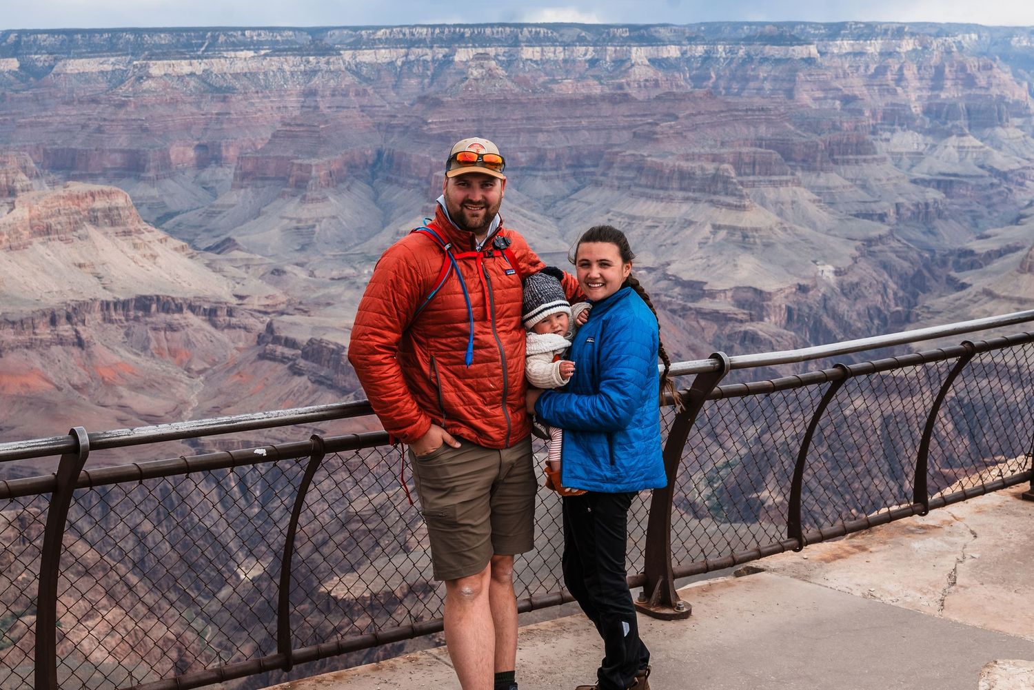 Visitors in bright orange and blue jackets stand at a scenic Grand Canyon viewpoint with protective railing on a cloudy day.