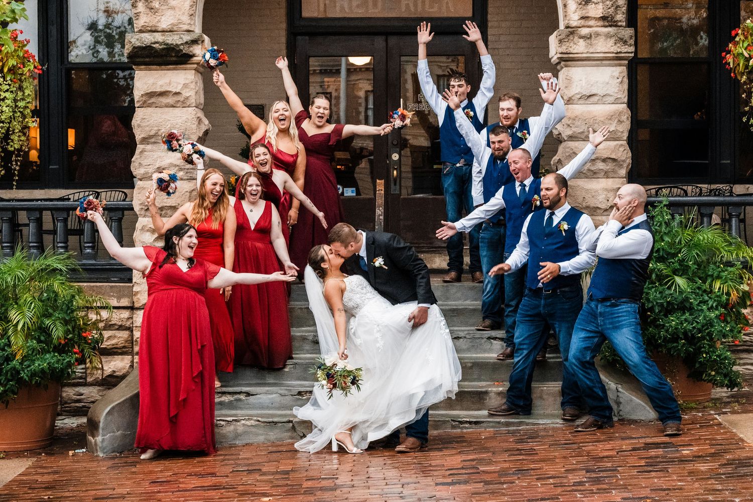 Wedding party celebrates with energetic jumping pose on brick steps of historic building.